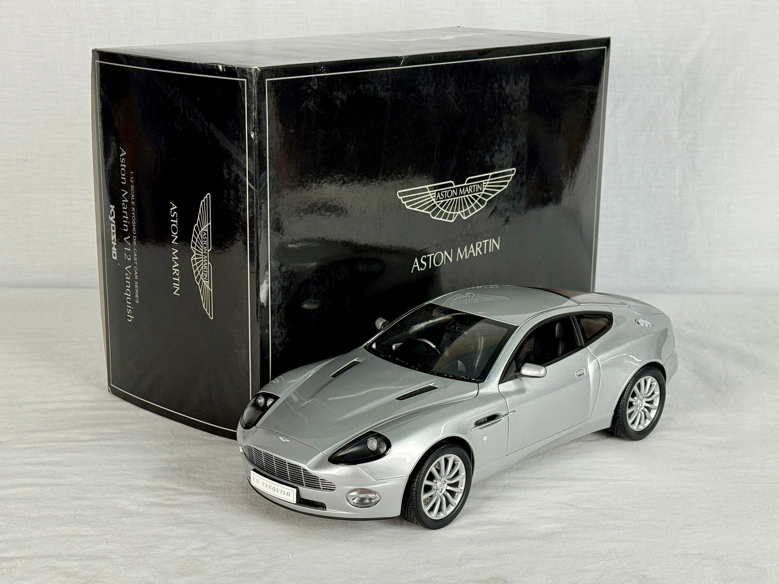 Kyosho 1:12 Scale Aston Martin V12 Vanquish Die-Cast Model: A silver Kyosho 1:12 scale die-cast model of an Aston Martin V12 Vanquish with its original black branded packaging, C9.