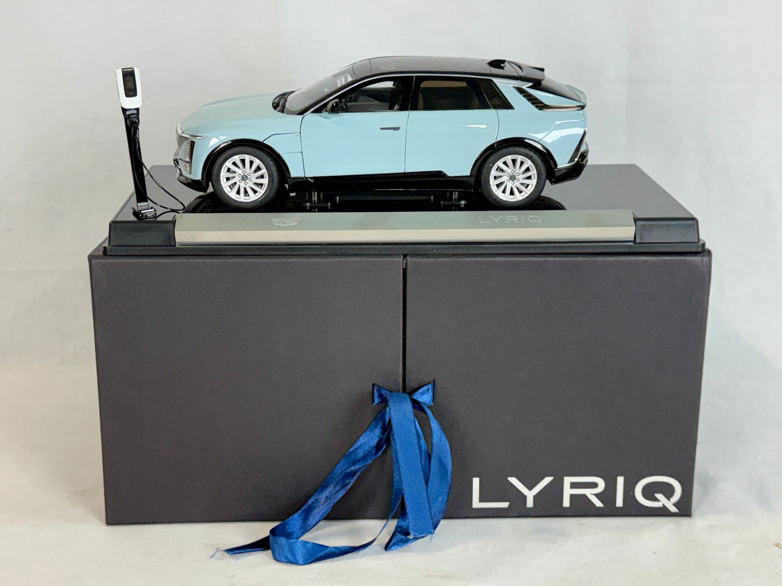 1:18 Scale Cadillac Lyriq Dealer Promo Model Car in Box: A 1:18 scale light blue Cadillac Lyriq electric SUV diecast model car featuring a black roof, mounted on a branded display base with a miniature charging station and original presentation box. Resin,