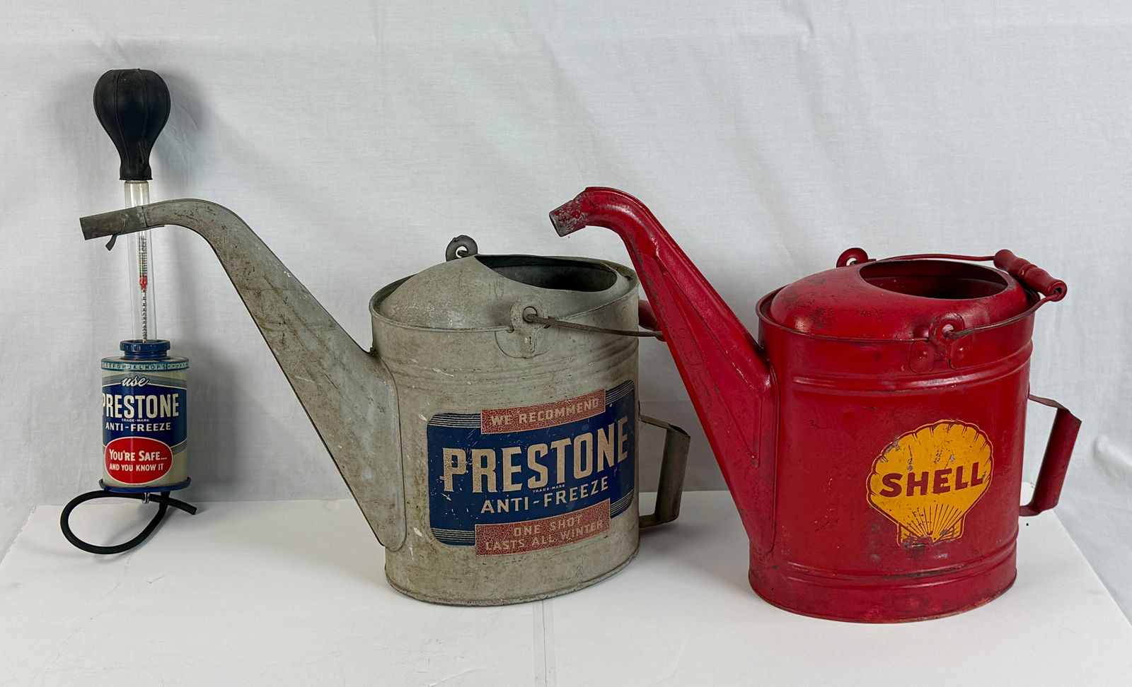 Three Vintage Prestone and Shell Automotive Service Items: A trio of vintage automotive service tools featuring a gray Prestone Anti-Freeze metal pour can, a red Shell logo metal pour can, and a Prestone anti-freeze tester with a rubber bulb, C5-7.
