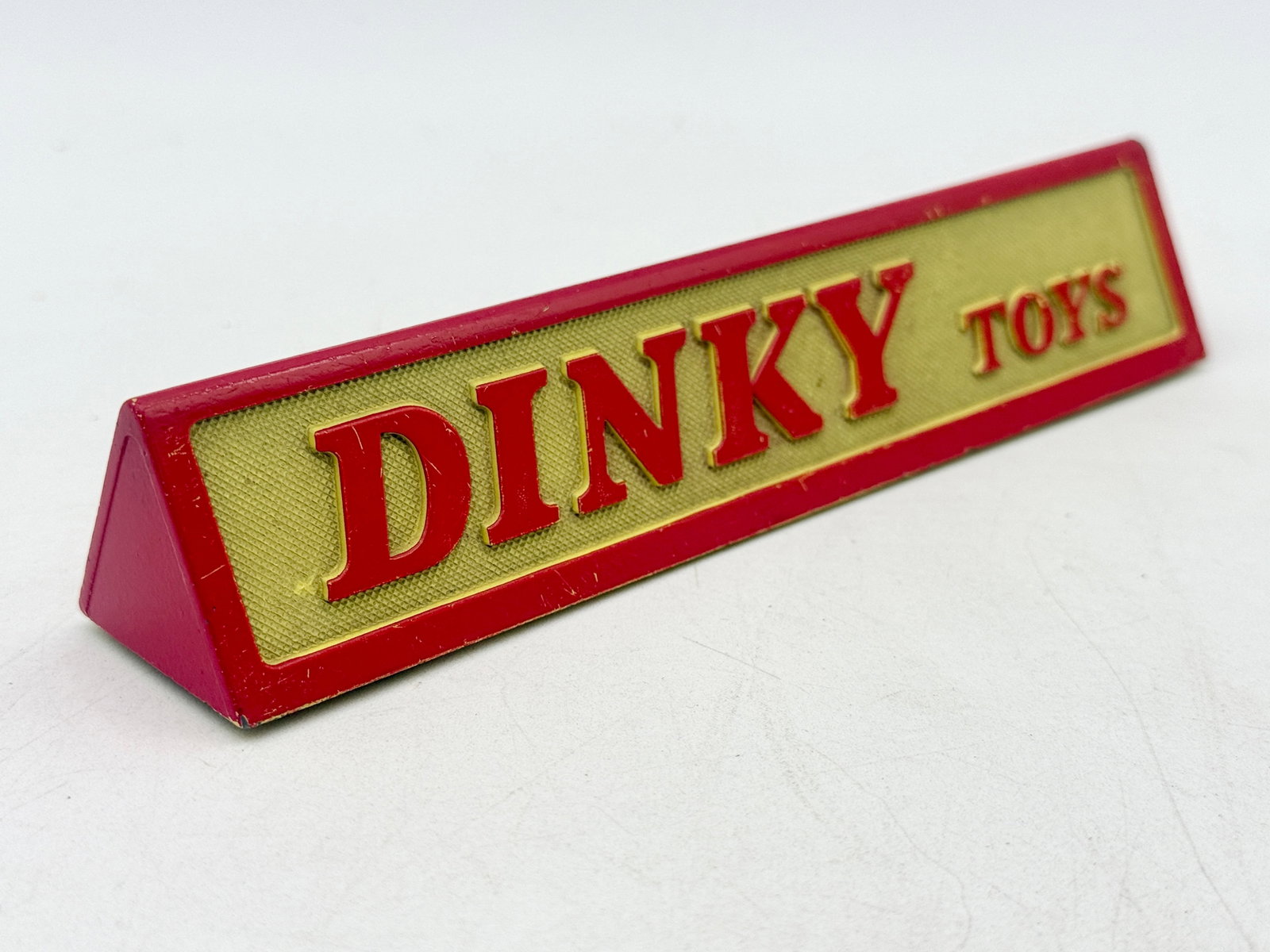 Vintage Dinky Toys Die-Cast Metal Triangular Shop Display Sign: This vintage shop display sign for Dinky Toys is constructed of die-cast metal in a triangular prism form. The front features a textured gold-toned panel with raised red lettering, while the hollow re
