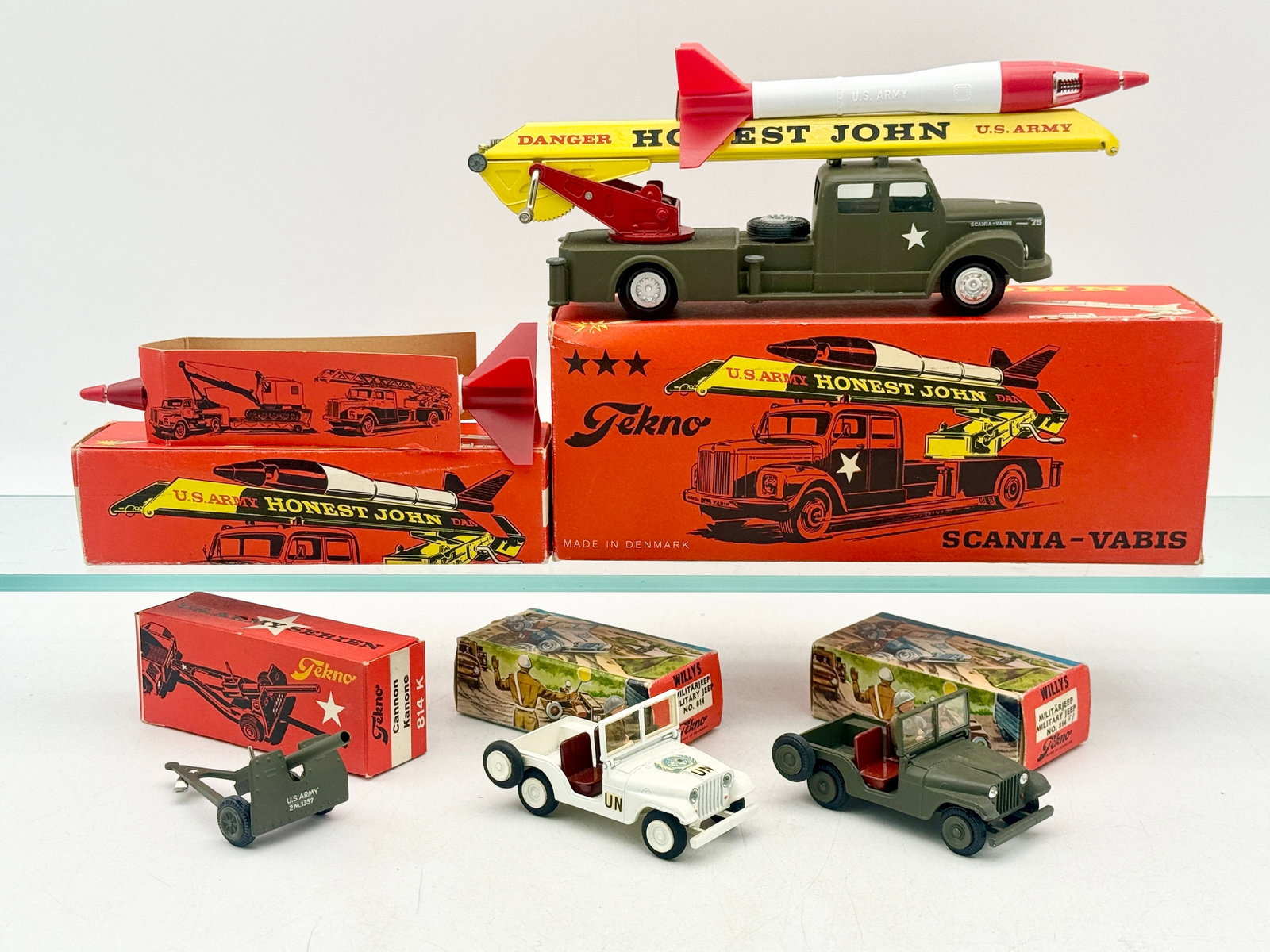 A Collection of Tekno Denmark Die-Cast Military Vehicles and Boxes: An assortment of Tekno Denmark die-cast military toys including a Scania-Vabis Honest John missile launcher, an additional missile, two Willys jeeps, and a field cannon. These models are constructed o