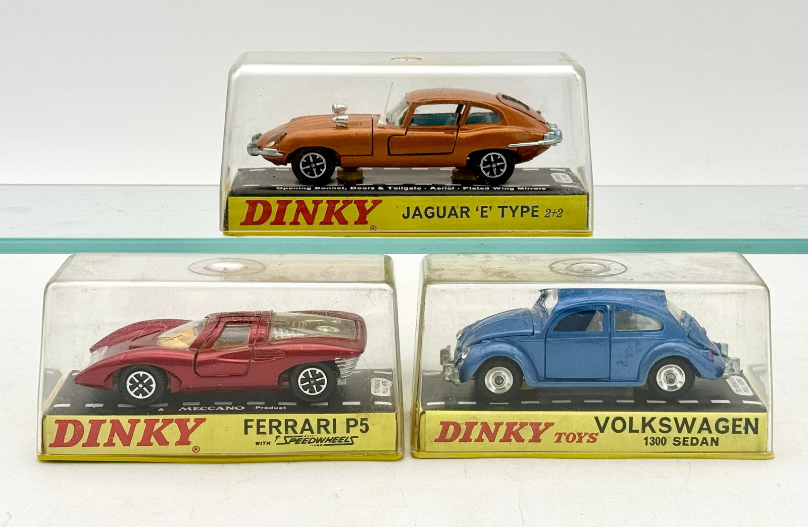 A Trio of Vintage Dinky Toys Die-Cast Model Cars: A trio of vintage Dinky Toys die-cast model cars featuring a metallic bronze Jaguar E-Type 2+2, a red Ferrari P5, and a blue Volkswagen 1300 Sedan. Each vehicle is constructed of die-cast metal and is