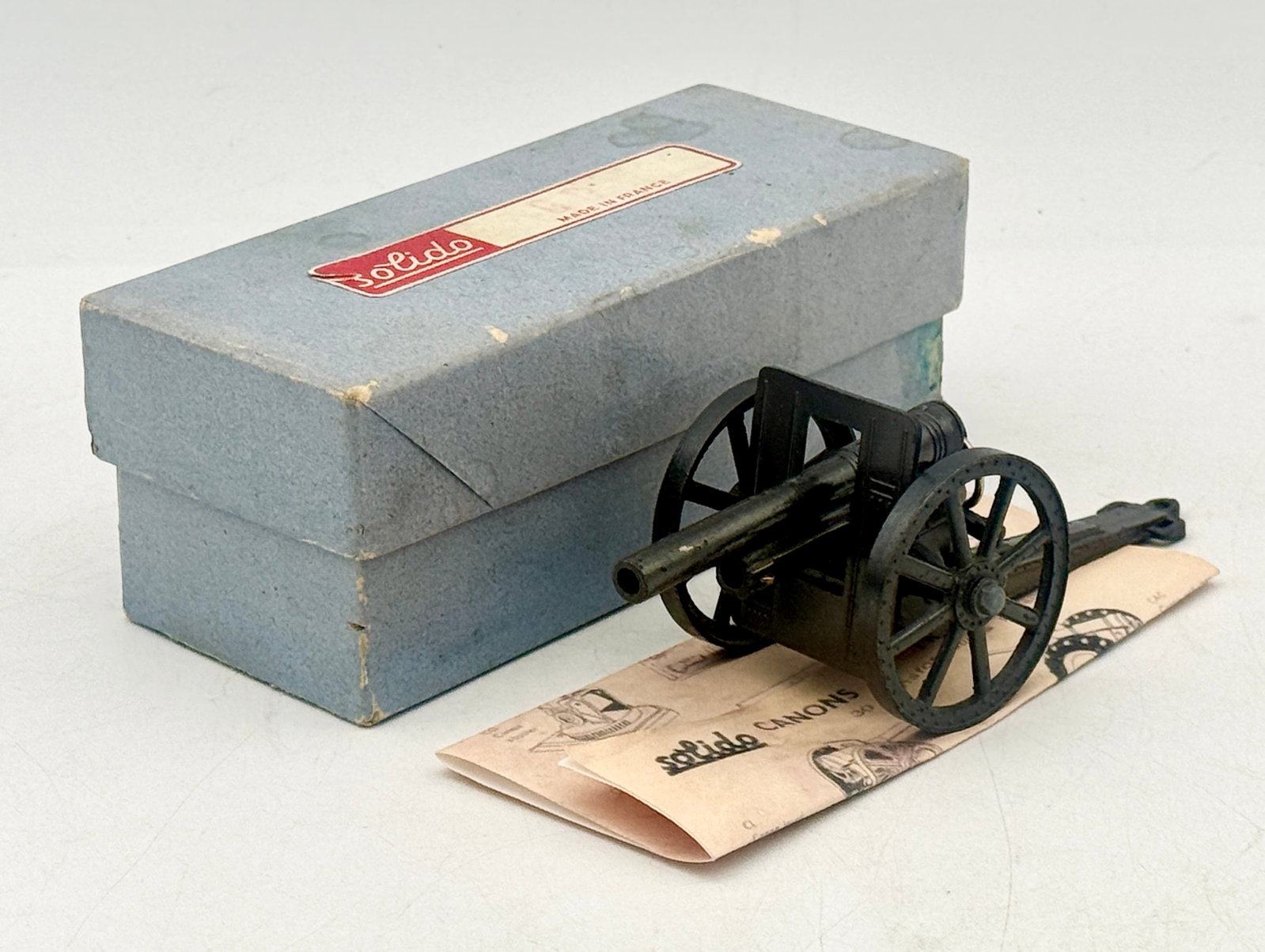 Solido Die-Cast Metal Toy Cannon with Original Box and Leaflet: A vintage Solido die-cast metal toy cannon produced in France, finished in a dark, aged patina. The model features spoked wheels, a protective gun shield, an adjustable elevation gear, and a spring-lo