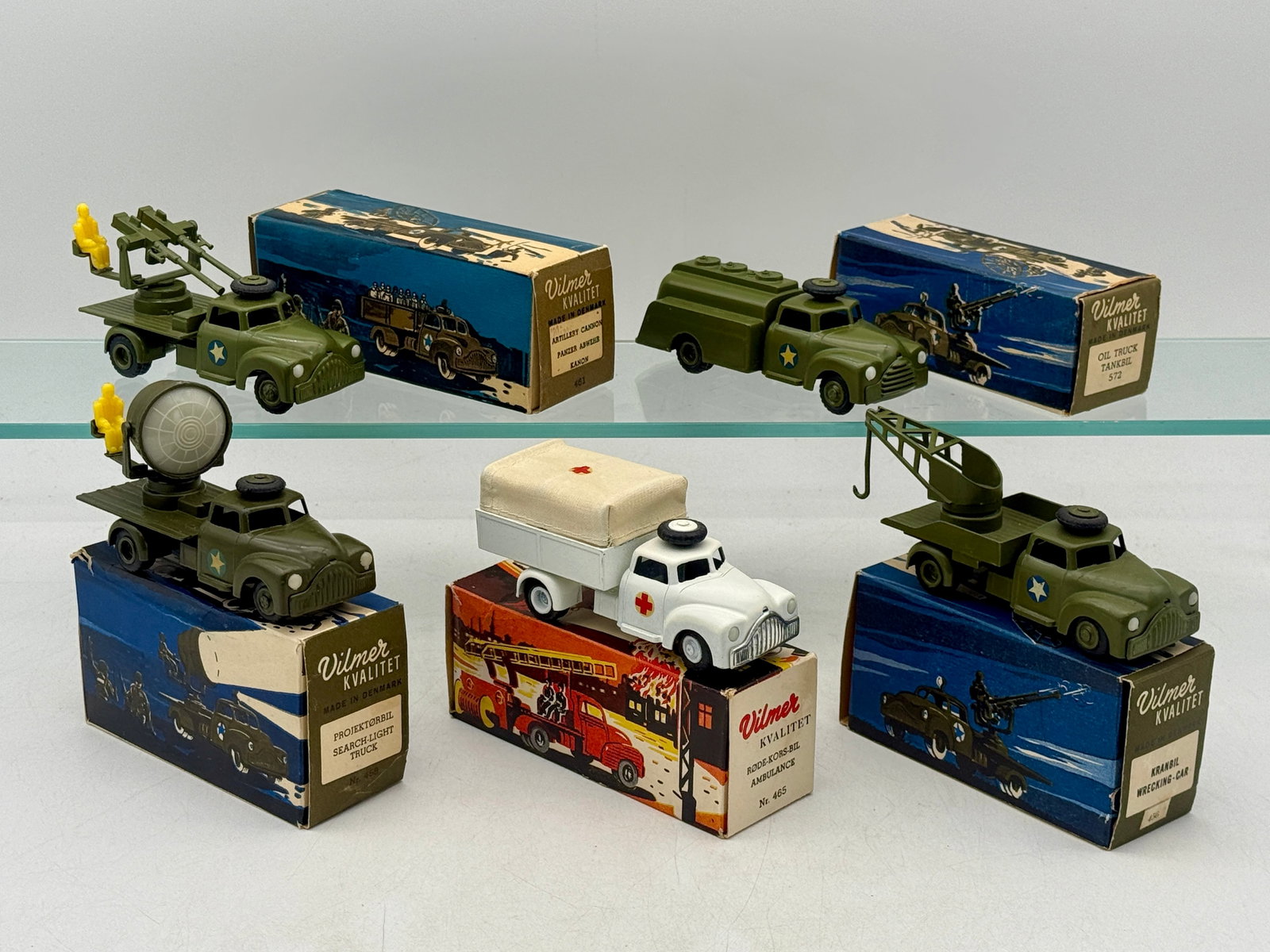 A Collection of Vintage Vilmer Die-Cast Military and Emergency Vehicles: This collection of Vilmer die-cast toy trucks, produced in Denmark, includes multiple military vehicles in olive drab and a white ambulance. Each vehicle is paired with its original pictorial cardboar