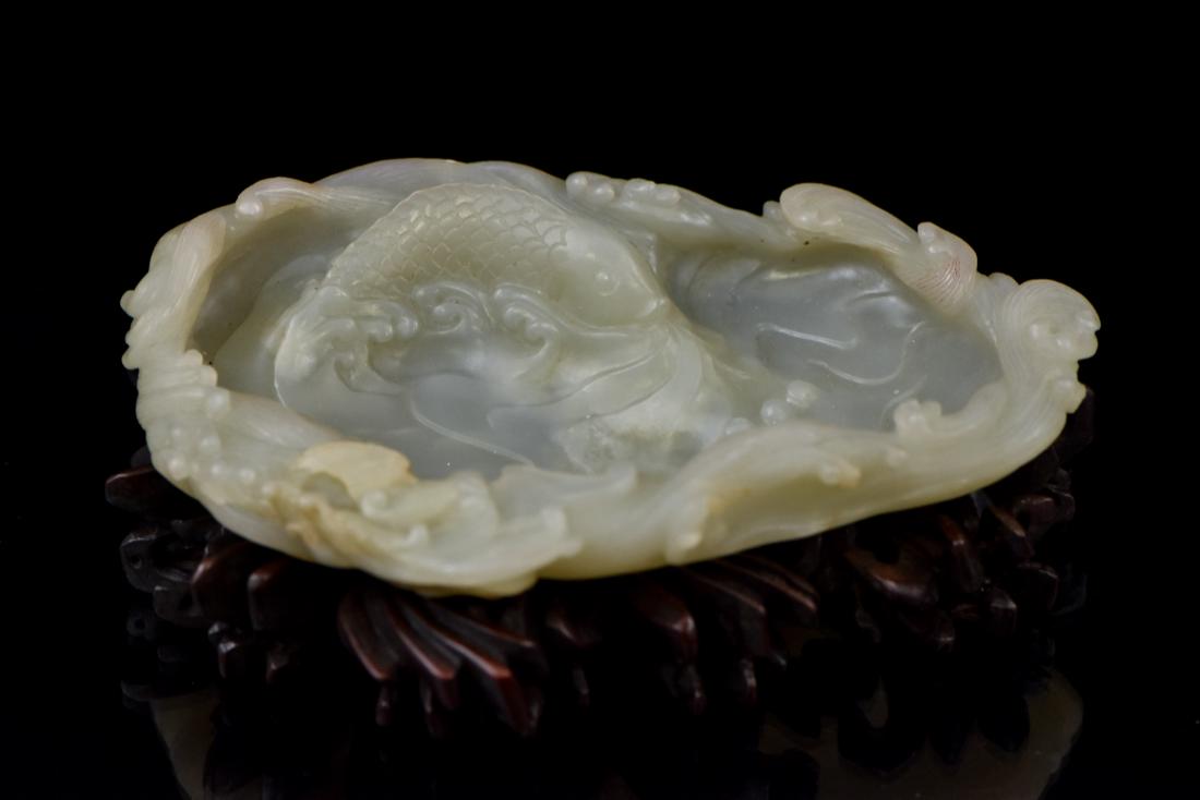 FISH ON POND MOTIF JADE BRUSH WASHER: A Chinese celadon jade carved in a shape of a shallow vessel with rim resemble a cresting waves on a pond. A carp emerged from a rippled water on the interior. The jade carving is superb, showing pale