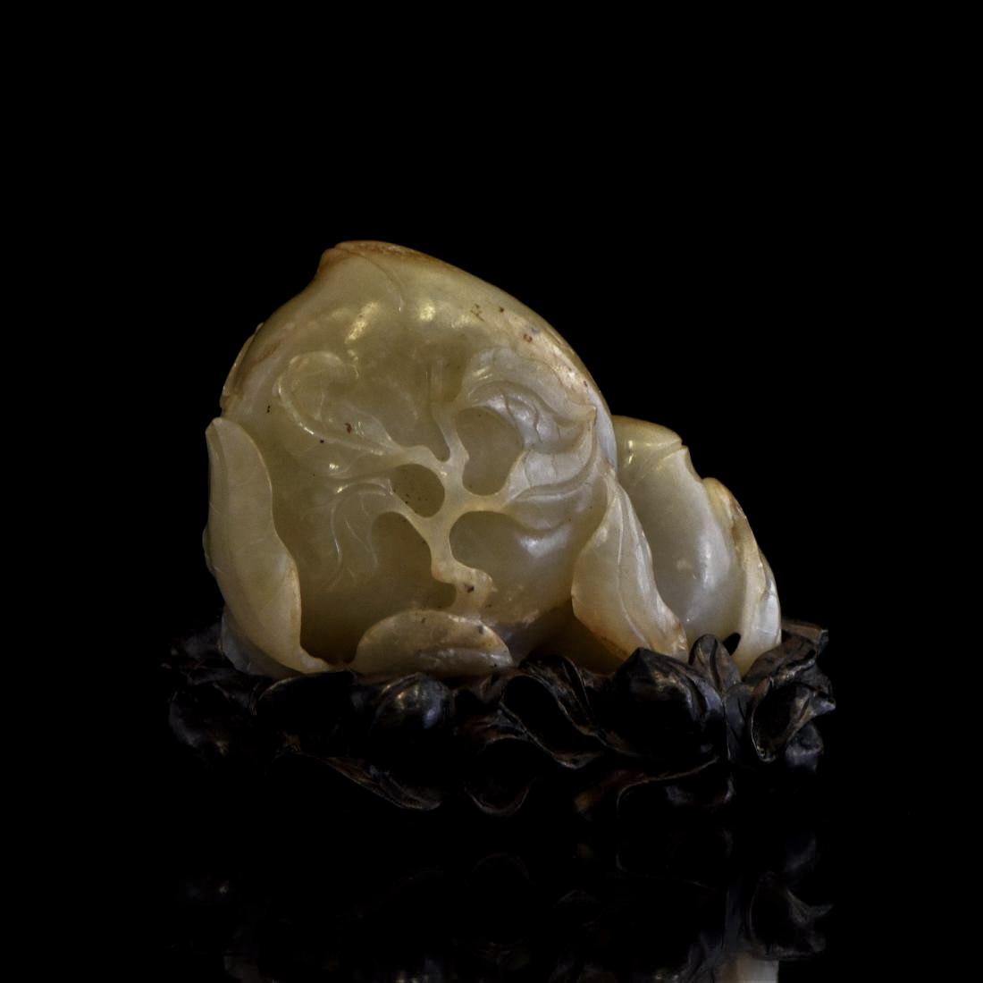 CARVED CREME-TEAL JADE OF PEACHES AND BAT ON STAND: A nephrite jade carved in recumbent two peaches with its dentil foliage, with a Fu-bat rested on top. The jade shows creme-grayish color, with ,moist milky appearance. Displayed upon a carved hardwood