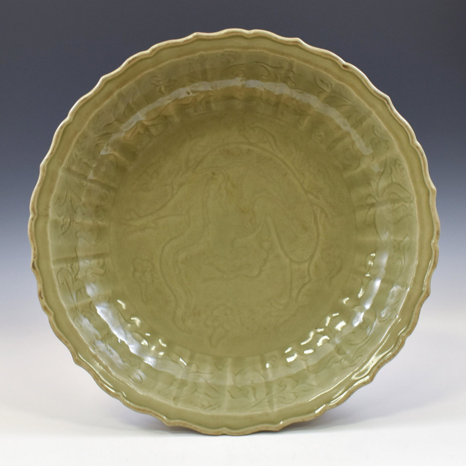 A LARGE CARVED DRAGON MING LONGQUAN CELADON CHARGER: Chinese Ming Dynasty LongQuan celadon barbed-rim charger. Shallow body, with a slightly flared lip with a shallow draft. Centrally festooned with a dynamic dragon soaring amidst flames and vapors. The