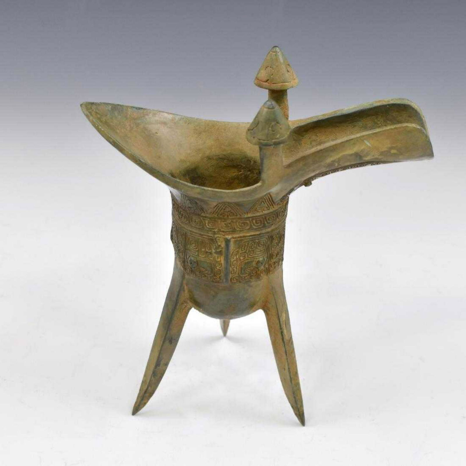 CHINESE ANTIQUE BRONZE JUE WINE VESSEL: Antique Chinese bronze ritual Jue wine vessel. The deep body is raised on three blade-shaped supports. Scored geometric patterns line the vessels outer diameter. Taotie mask is centered on a third fla