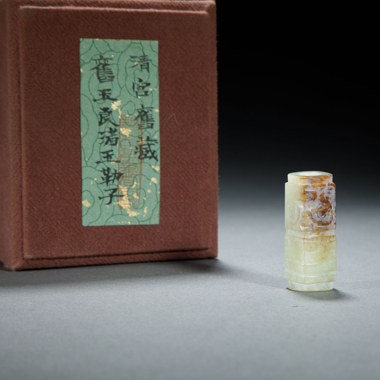 CHINESE HETIAN JADE LEZI BEAD: Dimensions: L. 1.5 cm. H. 4 cm. This elegant Chinese Hetian jade lezi bead features a cylindrical form, carved with archaic mask motifs in low relief. Its pale celadon stone is accented with