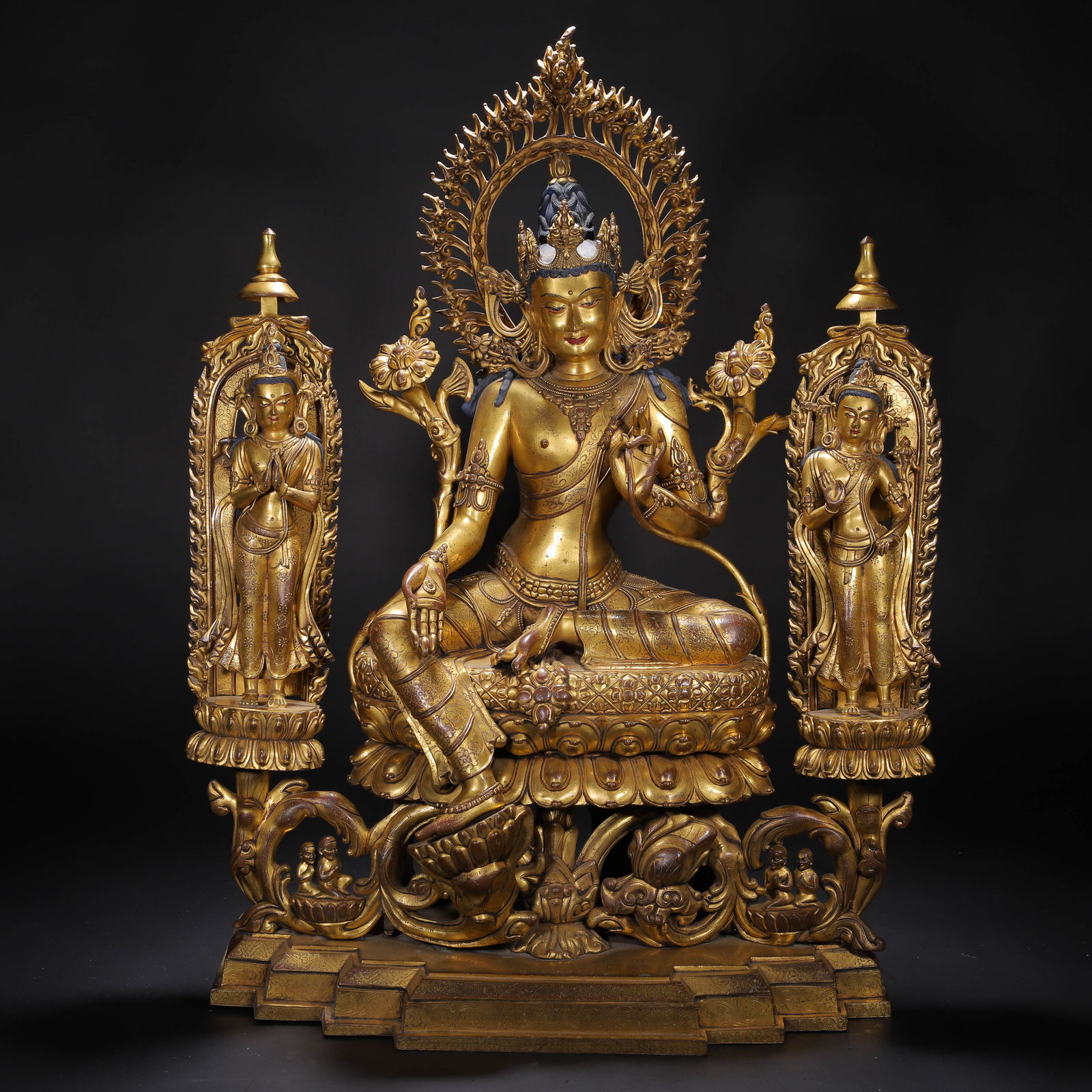 CHINESE GILT-BRONZE TRIAD: Dimensions: L. 56 cm. W. 27 cm. H. 73 cm. This imposing Chinese gilt-bronze triad centers on a seated bodhisattva, flanked by two standing attendants, all set within elaborate foliate aureoles. The ce