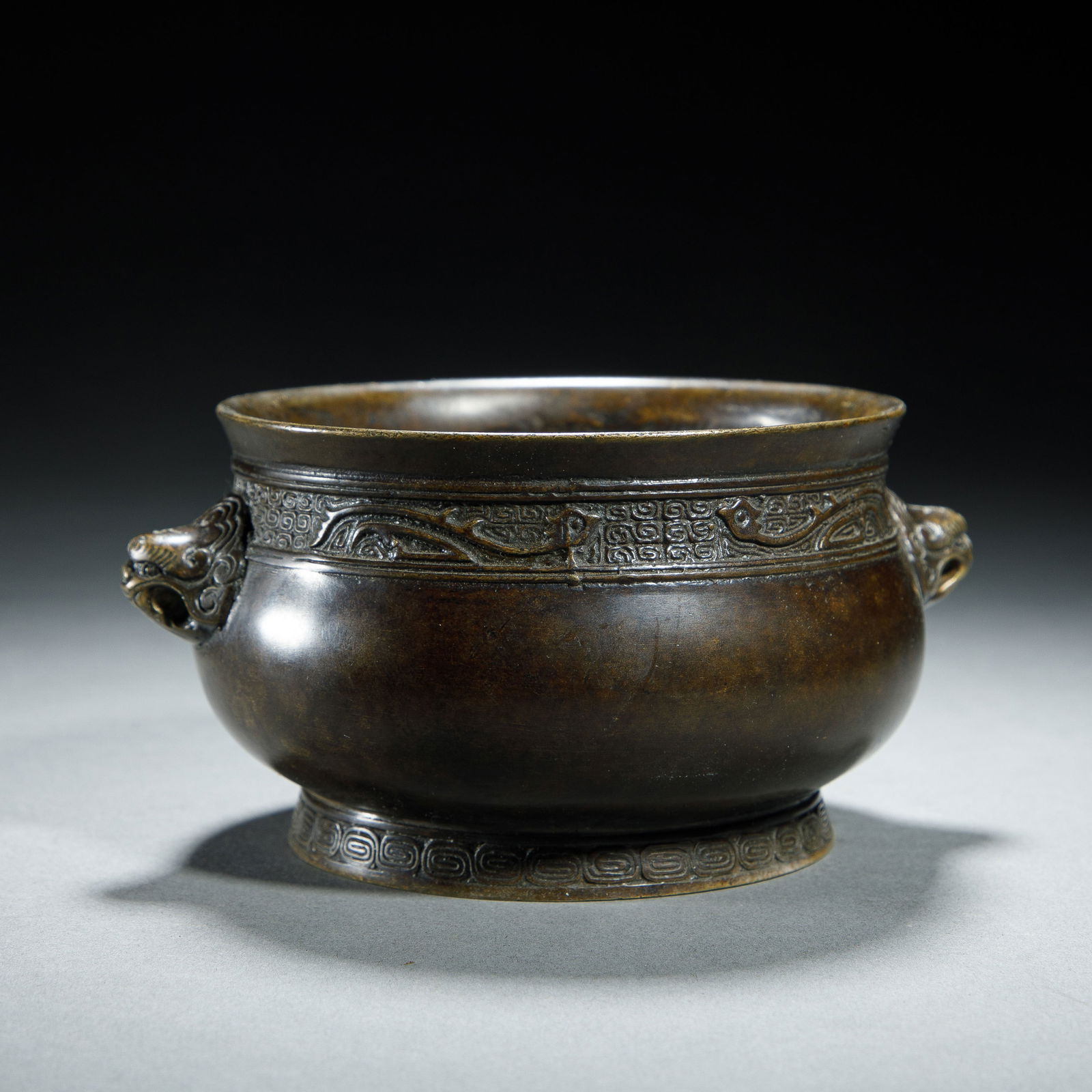 CHINESE BRONZE CENSER: Dimensions: L. 13.5 cm. H. 7 cm. This refined Chinese bronze censer features a rounded, bulbous body with a wide, everted rim, flanked by two relief lion-head handles. The neck is decorated with a ban