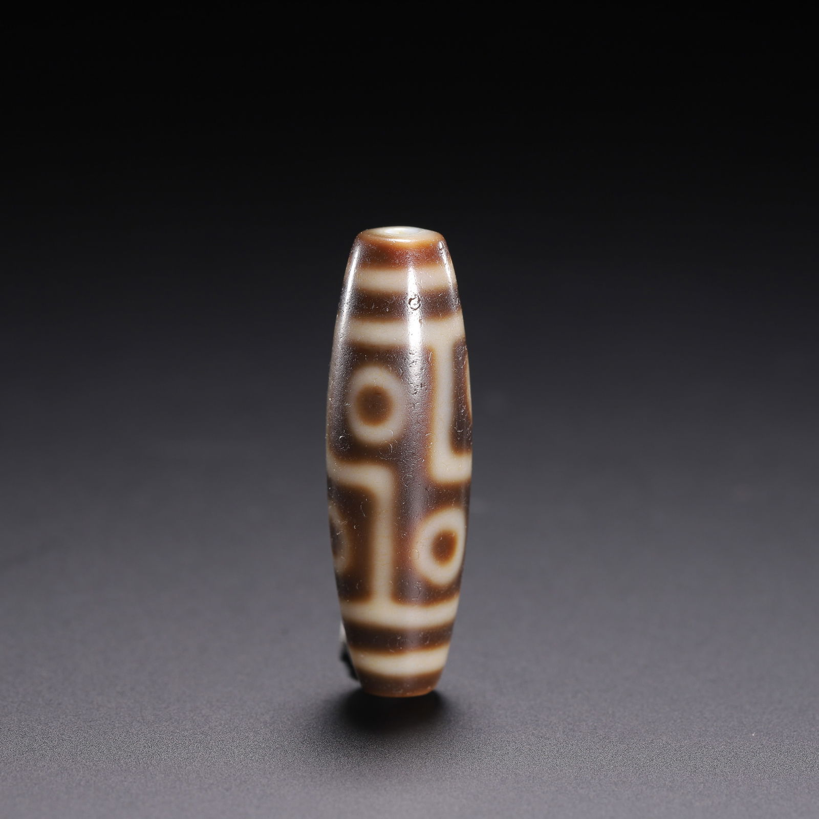 CHINESE DZI BEAD: Dimensions: L. 4 cm. W. 1.3 cm. Weight 10 grams. This striking Chinese dzi bead features a rich, chocolate-brown ground with crisp, creamy-white geometric patterns, including stylized eyes and