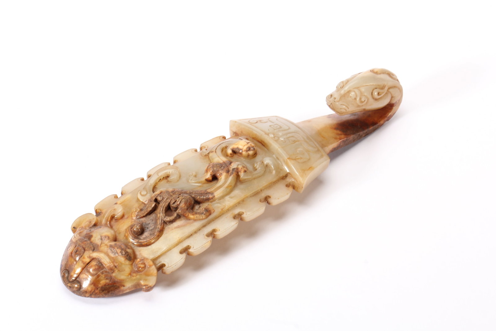 A CHINESE ARCHAIC CARVED JADE BELT HOOK: Weighing 170 grams, this elegant archaic belt hook measures 17.5 cm long, 4 cm wide and 3 cm tall. Crafted from pale nephrite marked by rich natural russet inclusions, it is topped by a finely rendere