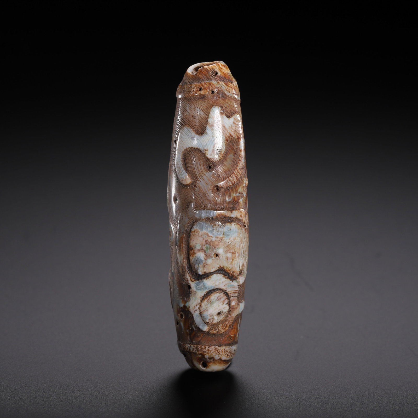 A CHINESE CARVED DZI BEAD: This elongated cylindrical dzi bead has a length of 7 cm and a total weight of 30 grams. Its textured surface features hand-carved stylized symbolic motifs contrasting against mottled earthy brown, cr
