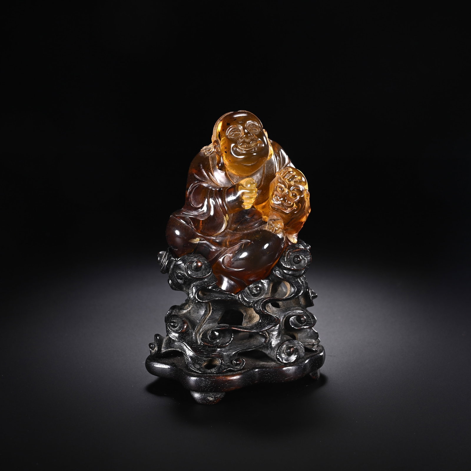 A CHINESE AMBER LUOHAN ORNAMENT: Standing 10 cm tall and 6.5 cm wide, this charming sculptural piece is carved from translucent warm amber showcasing natural honey-toned tonal gradients and attractive light refraction. The seated Luo