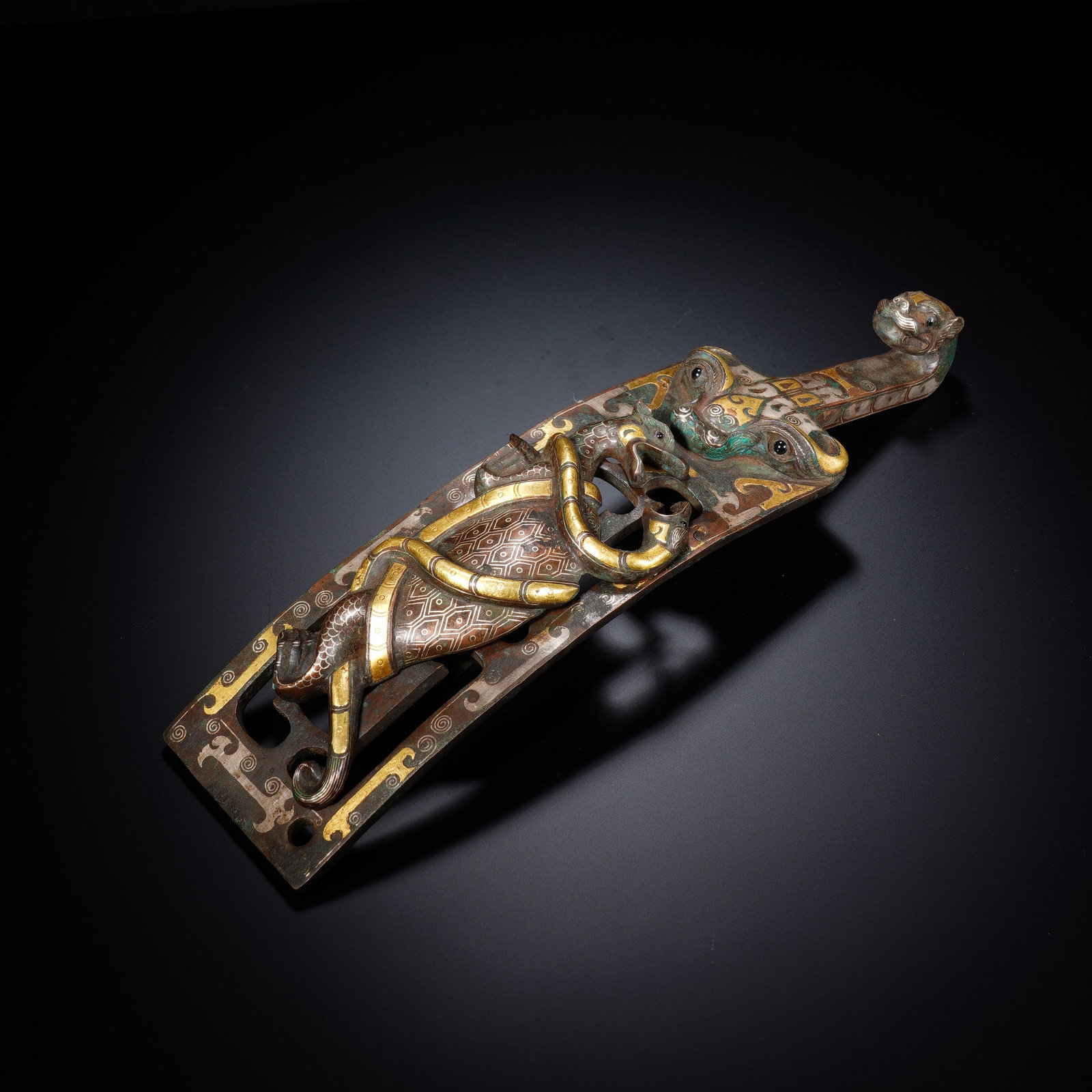 A CHINESE INLAID BRONZE BELT HOOK: Fashioned from solid bronze, this striking belt hook features masterful multi-metal inlay work with gleaming gold and silver detailing across its elongated curved body. Elaborate openwork carvings sho
