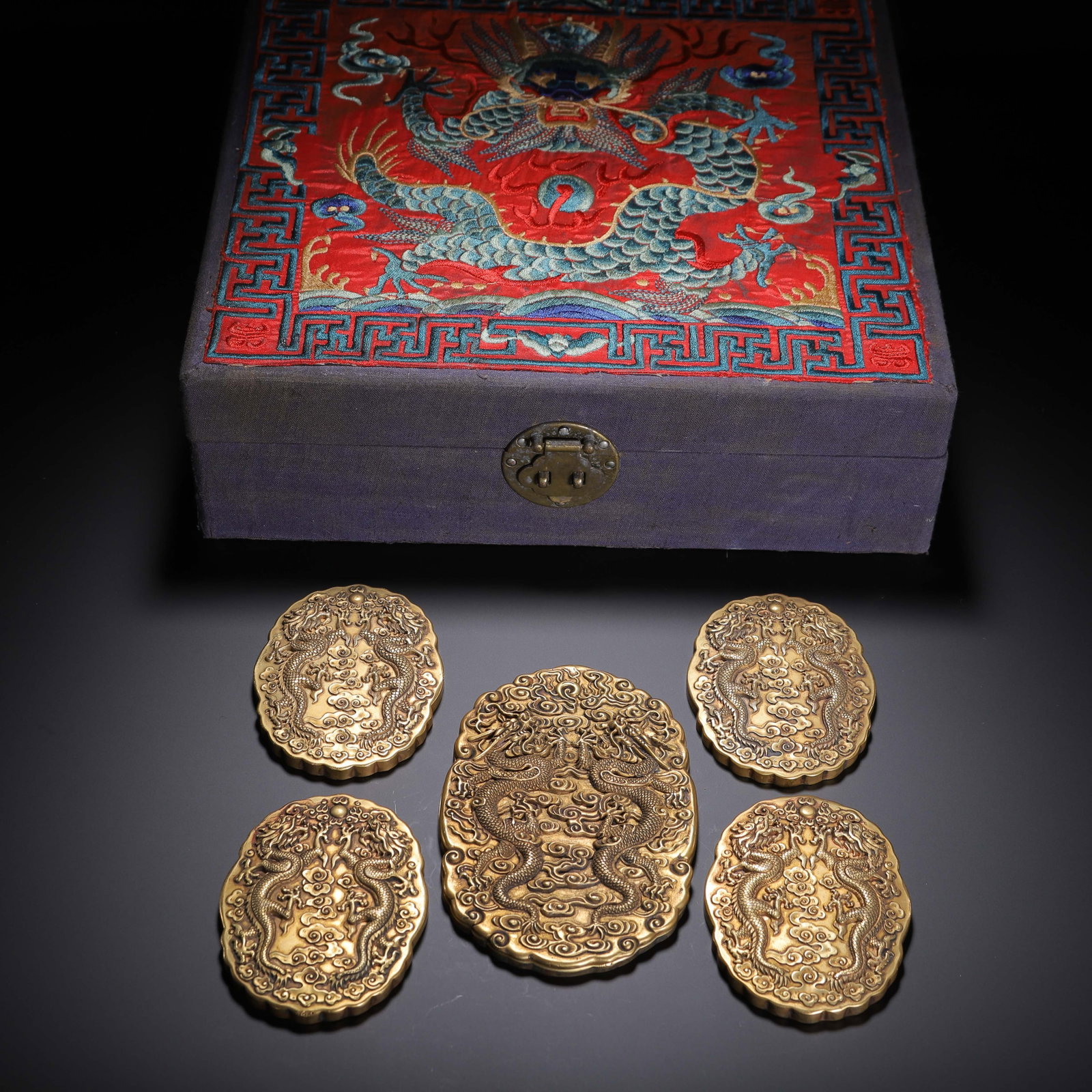 A CHINESE SET OF GILT BRONZE TOKENS: This elegant assemblage includes five elaborately cast gilt bronze tokens housed within a bespoke storage box. The box lid showcases vivid needlework: a prominent embroidered dragon unfurls across sca
