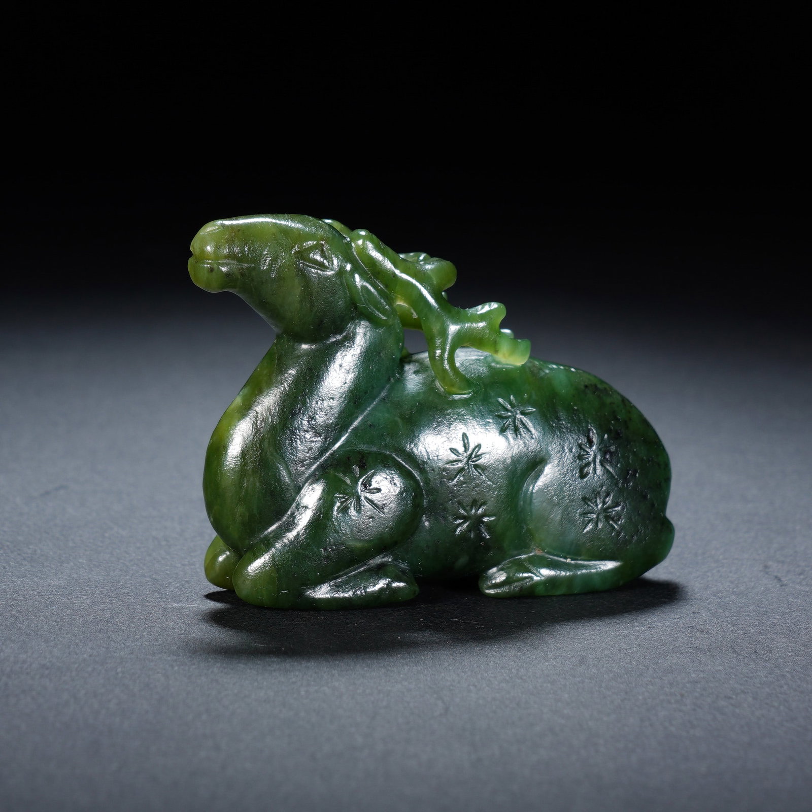 A CHINESE SPINACH JADE DEER FIGURE: Expertly carved from rich deep green spinach jade, this delightful ornament portrays a deer resting gracefully in a recumbent pose with elegantly shaped branching antlers. Delicate incised star-shaped