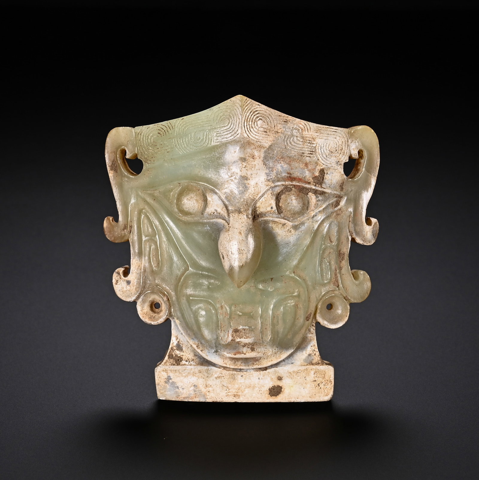 A CHINESE HETIAN JADE MASK ORNAMENT: Measuring 6.3 cm tall and 5.6 cm wide, this finely carved mask is fashioned from pale celadon Hetian jade mottled with natural creamy earthy patination across its lower edges and flat rectangular plin