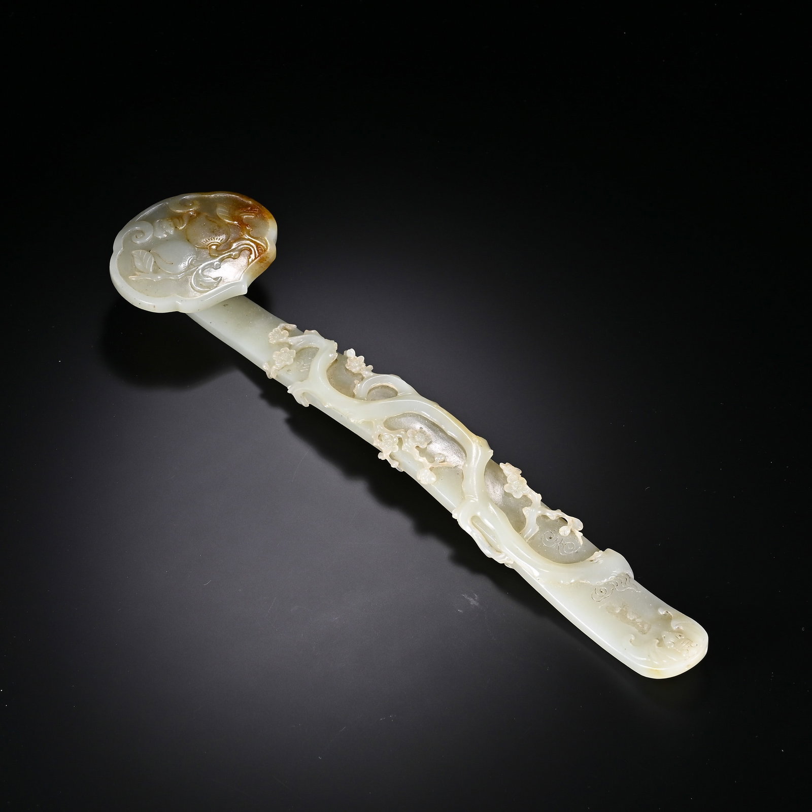 A CHINESE HETIAN JADE RUYI SCEPTRE: Masterfully carved from fine Hetian jade with a creamy pale base, this elegant ruyi sceptre features natural warm russet patination prominently spread across its rounded scalloped head. Slender curvin