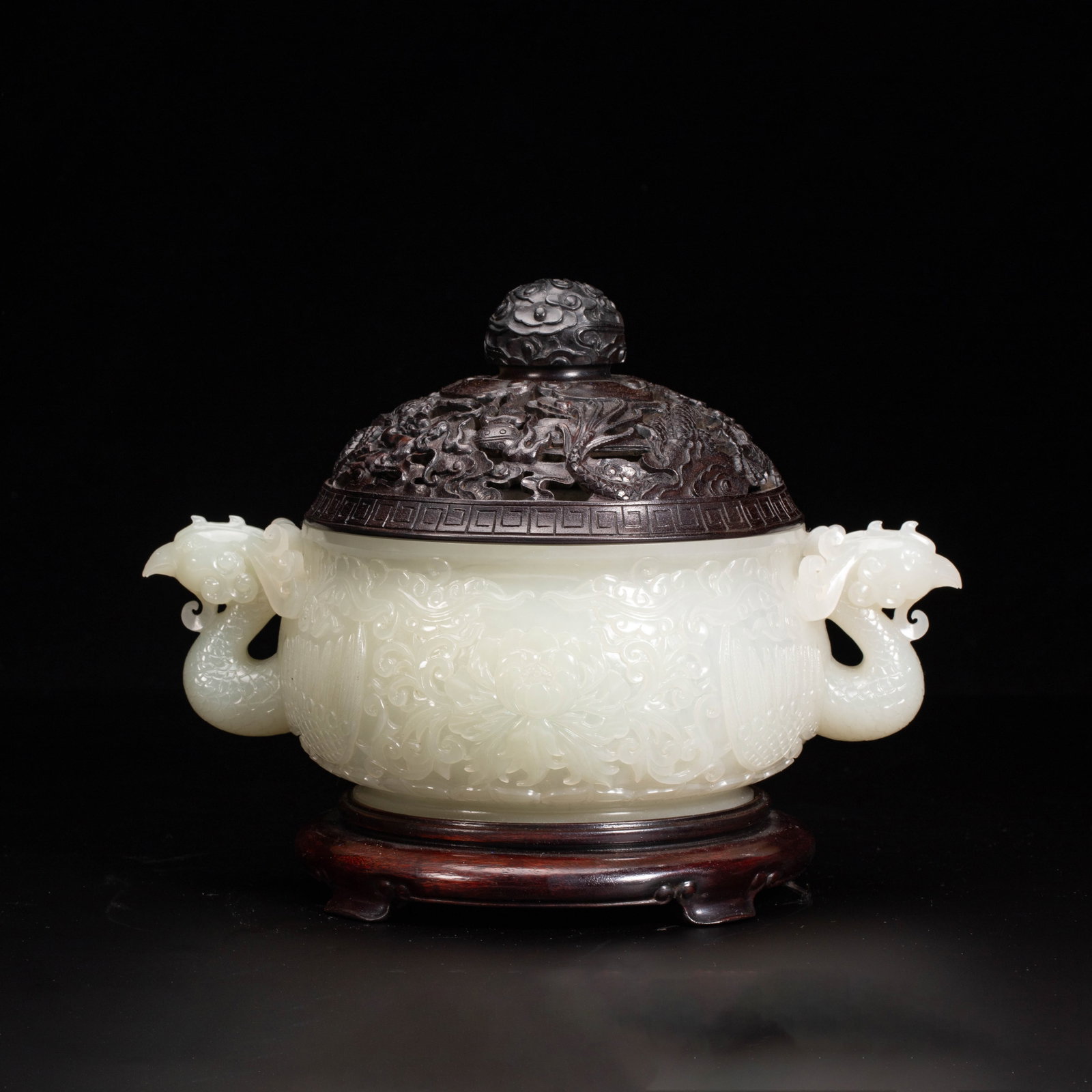 A CHINESE HETIAN JADE CENSER: Crafted from luminous creamy Hetian jade, the main vessel of this censer is adorned with deeply rendered floral scroll carvings across its rounded body. Symmetrical phoenix-head handles extend gracefu