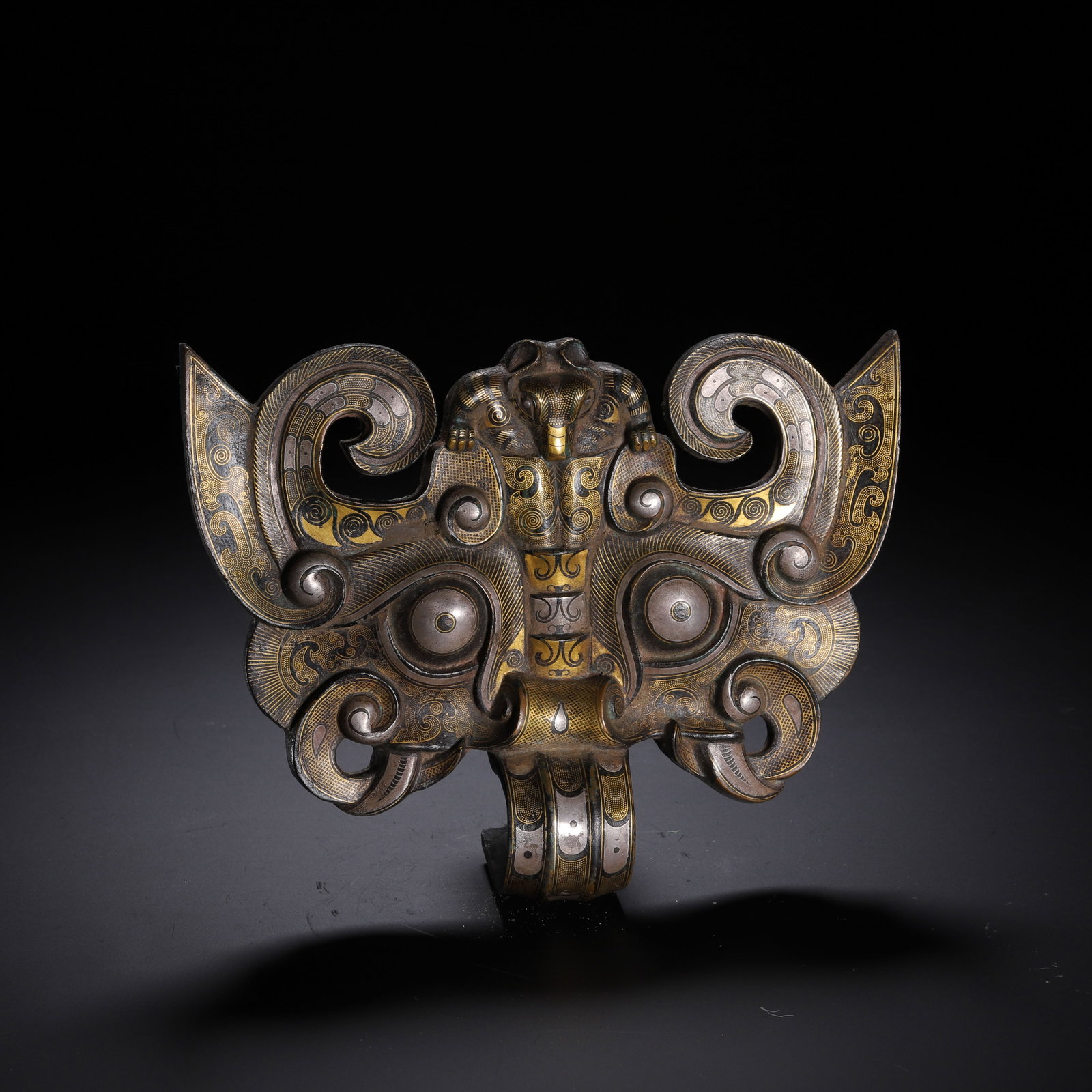 A CHINESE GOLD SILVER INLAID BRONZE TAOTIE FITTING: This striking bronze ornament is meticulously decorated with interwoven gold and silver inlay, fashioned as a formidable taotie beast mask with dramatically curled, flared aurals. Bulbous inset eyes d