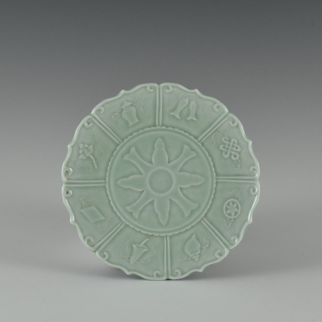 SGRAFIATTO PALE-CELADON BAJIXIANG FLORIFORM RIM PLATE: Stoutly potted unto a shallow vessel with gently rounded sides terminating the flori-form lobed of eight upward petal-barbed rim, each petal enclosed the bajixiang Buddhist emblems. The central interi