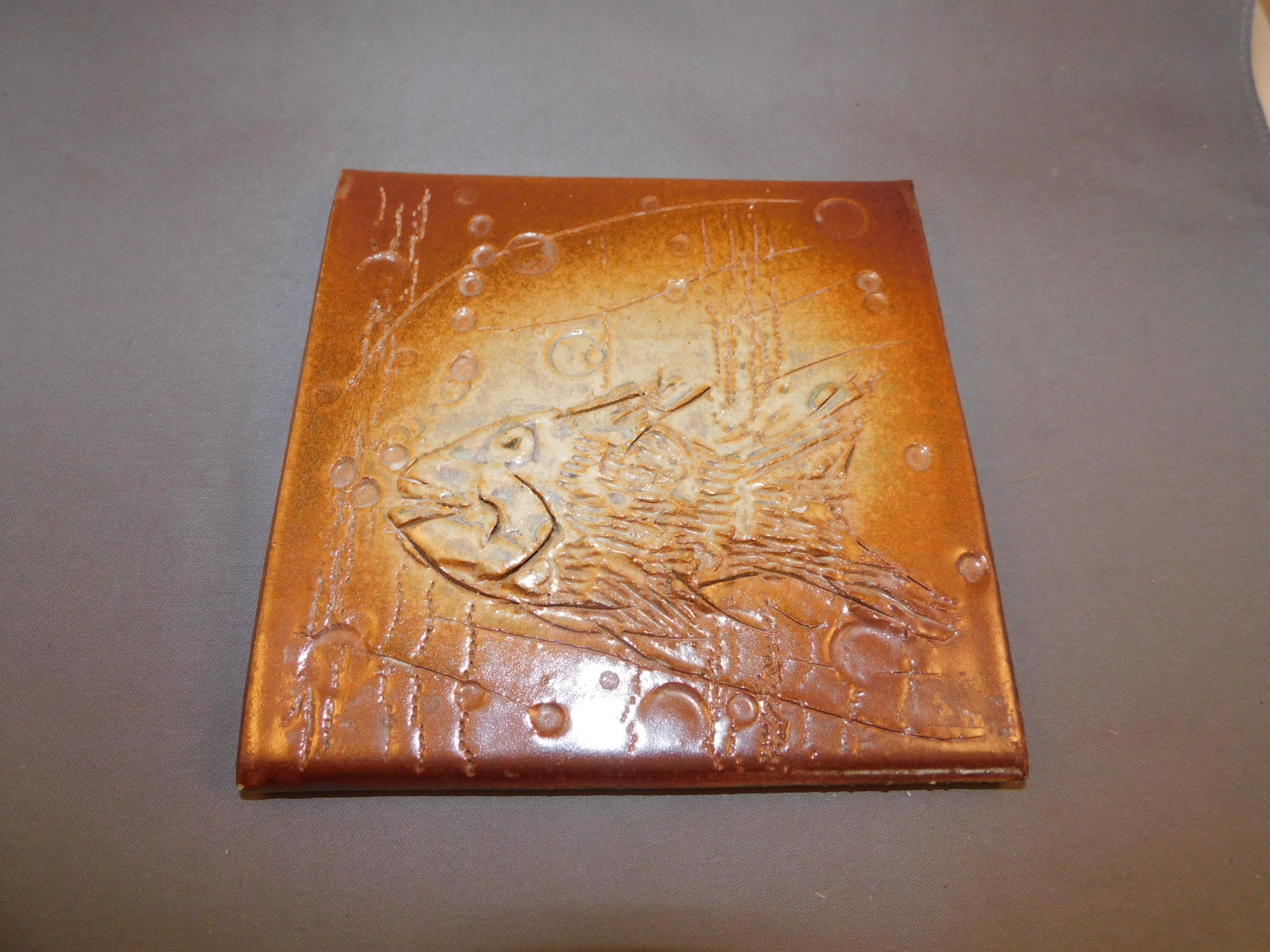 Art Pottery Fish Tile: Art pottery tile with a 3D fish on the front and bubbles throughout. Measures approx. 6" x 6". You are bidding at a live sale. Please read the terms and conditions. We are offering contactless pick up