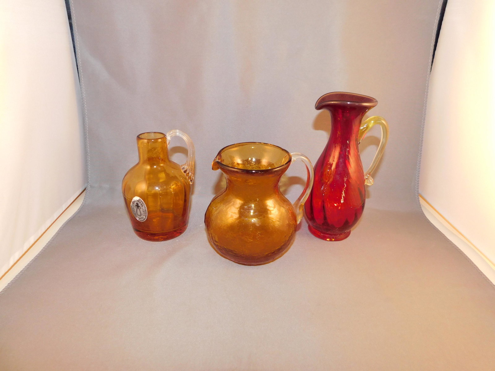 Lot of 3 Miniature Pitchers: Lot of 3 miniature pitchers including hand blown pilgrim glass and cranberry applied handled pitcher. Measures approx. 3 1/2 - 5 1/2" tall x 2" - 3" wide. You are bidding at a live sale. Please read t