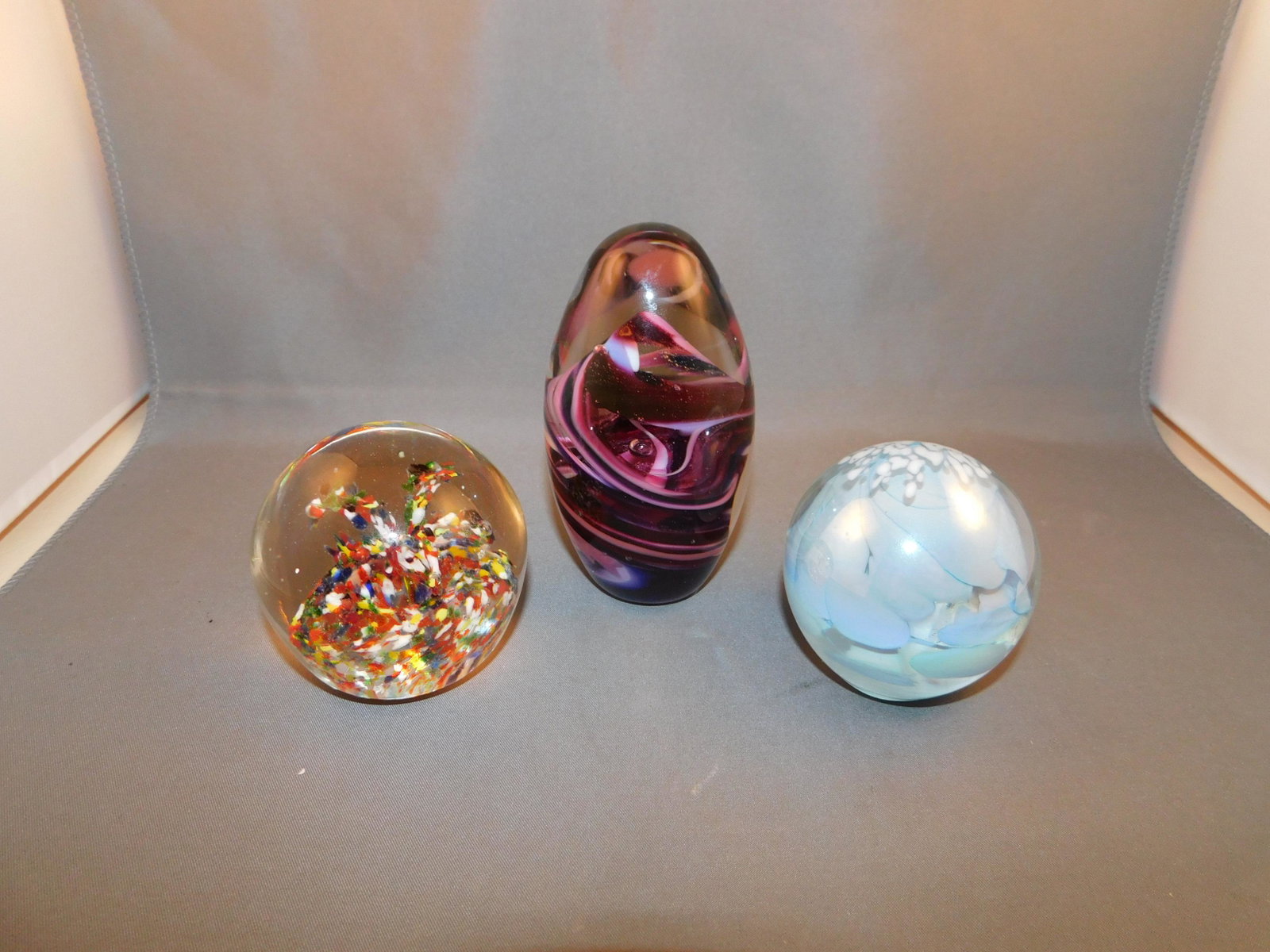 Lot of 3 Art Glass Paperweights: Lot of 3 art glass paperweights including a round multi colored, a blue iridescent and an purple swirl egg. Measures approx. 2 1/2" - 4" tall x 2" - 2 1/4" wide. You are bidding at a live sale. Please