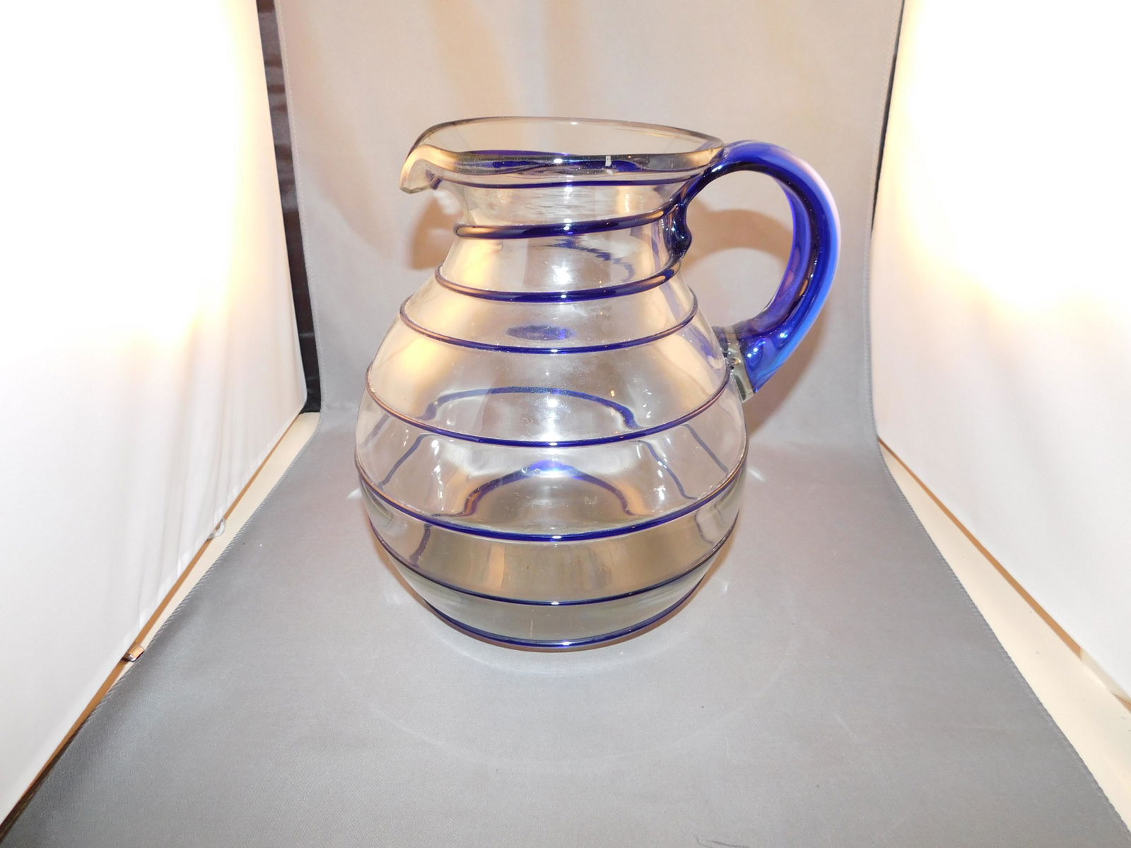 Art Glass Pitcher: Hand blown art glass pitcher with a blue glass swirl and blue glass applied handle. Measures approx. 8 1/2" tall x 8" wide. You are bidding at a live sale. Please read the terms and conditions. We are