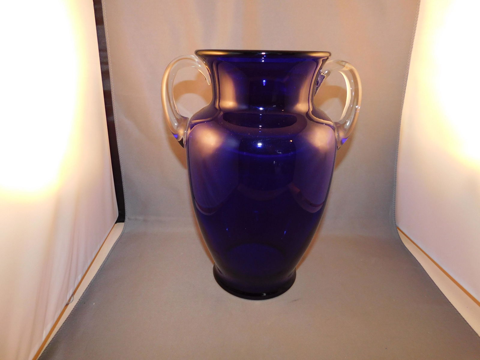 Art Glass Vase: Art glass double handled vase in cobalt blue with clear applied handles. Measures approx. 9 1/2" tall x 7" wide. You are bidding at a live sale. Please read the terms and conditions. We are offering n