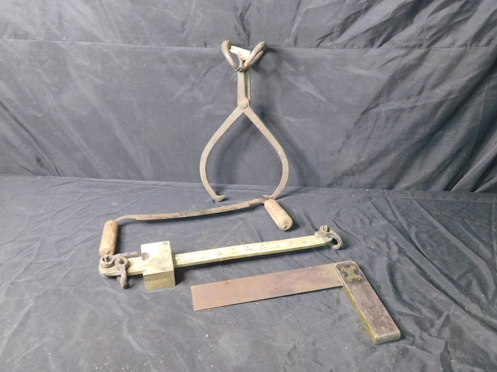 Primitive Tool Grouping: Vintage Primitive Tool Lot. Included is an Ice Pick, Brass Balance Scale Part, Primive Square with Brass, and a Double Handled Saw. This sale will be broadcast live on sale day with an auctioneer and