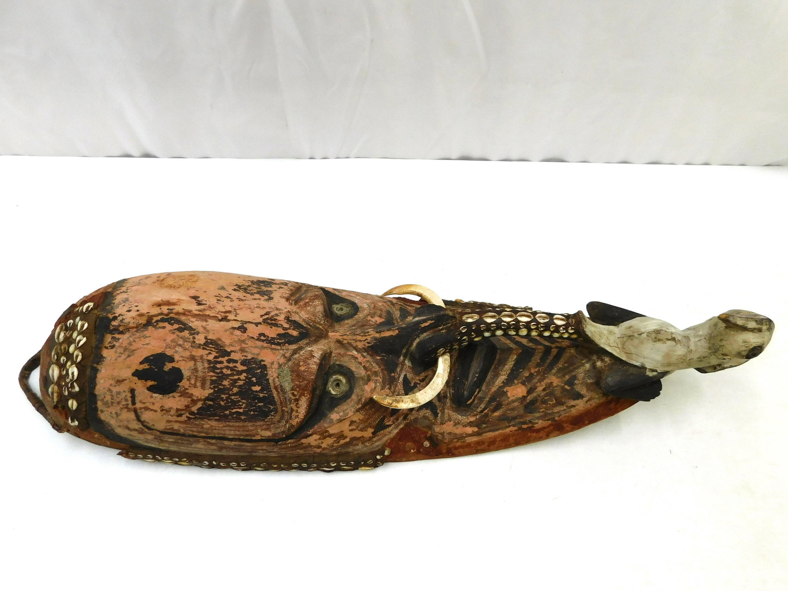 African Tribal Art Mask: African Tribal Art Mask. Hand Painted with Cowrie Shell Decoration and tusk. In the manner of Papua New Guinea. Has missing mud decoration throughout. Missing some shells. Measures 34" tall x 10" wide