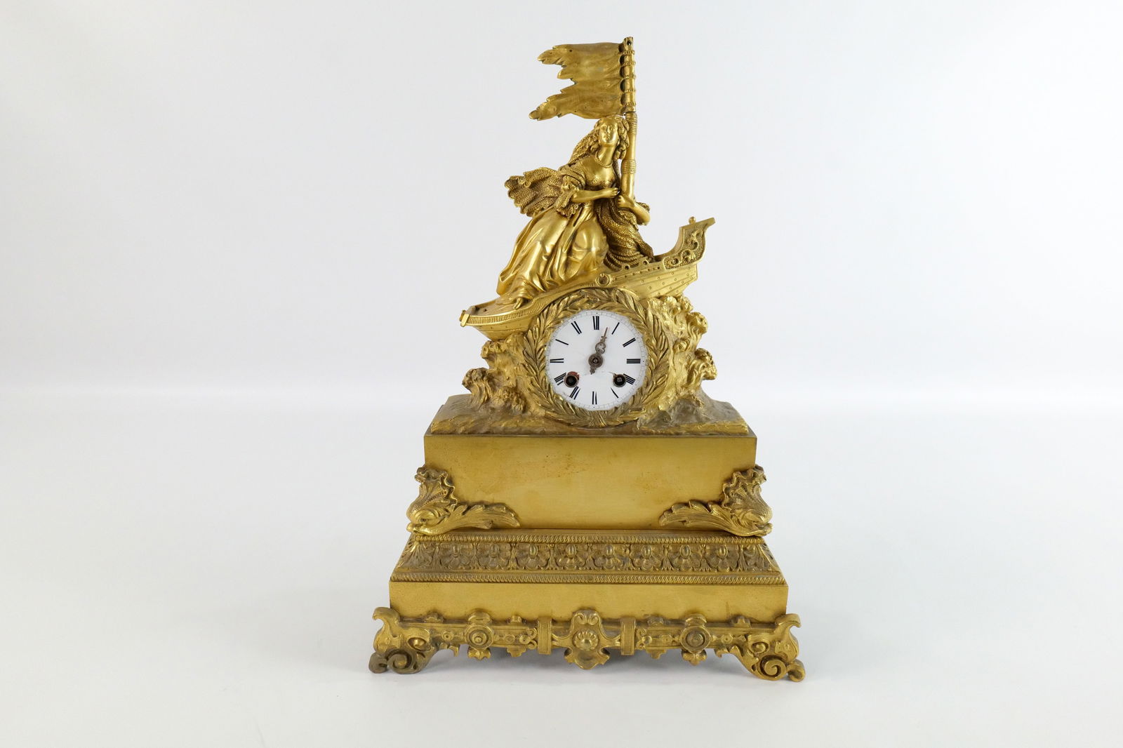 19th Century Bronze Mantle Clock: 19th Century Bronze Mantle Clock. Top crest is woman holding flag on a ship. Clock face has loss to porcelain and missing a hand. Works have some rusting. No Pendulum. Most likely French. Measures 14"