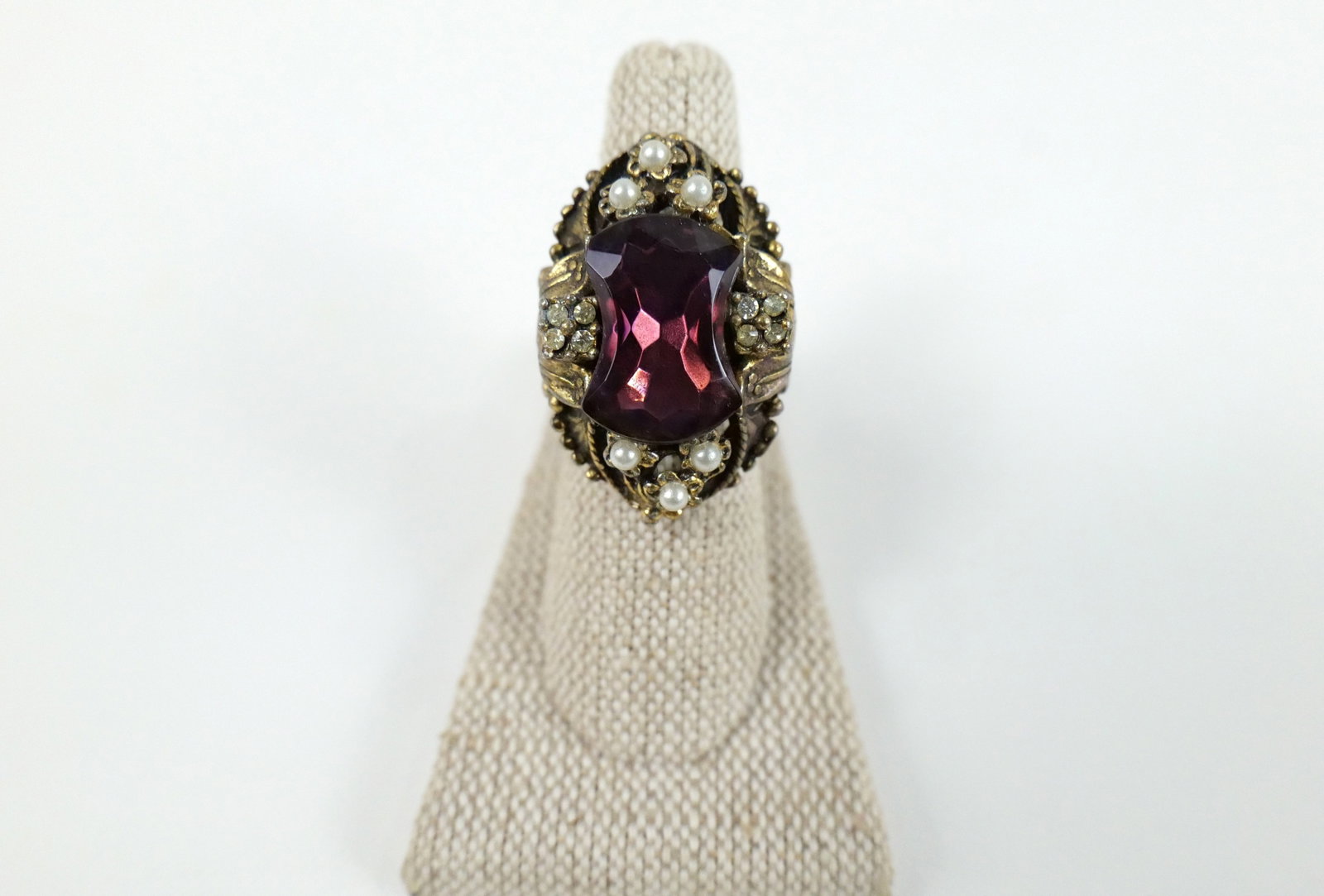 Vintage Gold Tone Cocktail Ring: Vintage Gold Tone Cocktail Ring. The lot includes 1 vintage c1970 adjustable gold tone cocktail ring measuring 1.33'' x .90'' and featuring a unique bow shaped purple glass gem in a floral setting wit