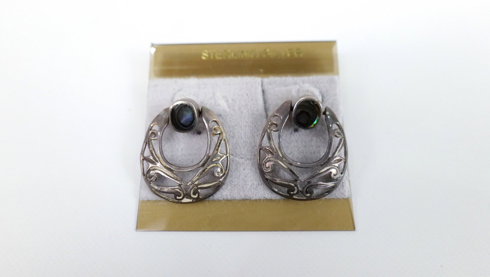 Carded Sterling and Abalone Earrings: Carded Sterling and Abalone Earrings. The lot includes 1 pair of vintage carded sterling front facing hinged hoop earrings measuring 1'' x .85'' and featuring a pierced scroll work motif with oval stu