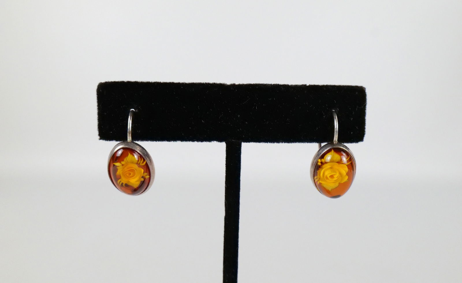 Reverse Carved Amber and Sterling Earrings: Reverse Carved Amber and Sterling Earrings. The lot includes a pair of .83'' x .43'' sterling framed earrings featuring 14mm x 10mm reverse carved amber cabochons in the image of a rose. They have hoo