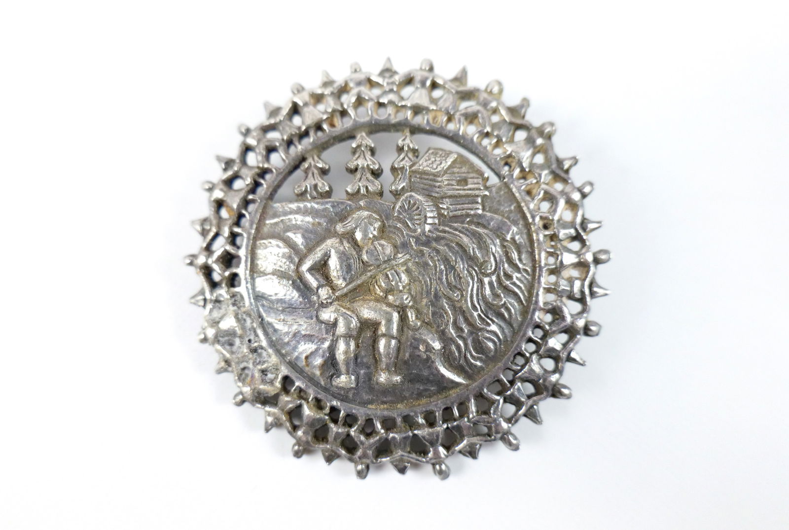 Pewter Holthe Tinn Norwegian Brooch: Pewter Holthe Tinn Norwegian Brooch. The lot includes a vintage 2.5'' Norwegian made pewter brooch depicting a man playing the fiddle next to a stream with an outer border of people holding hands and