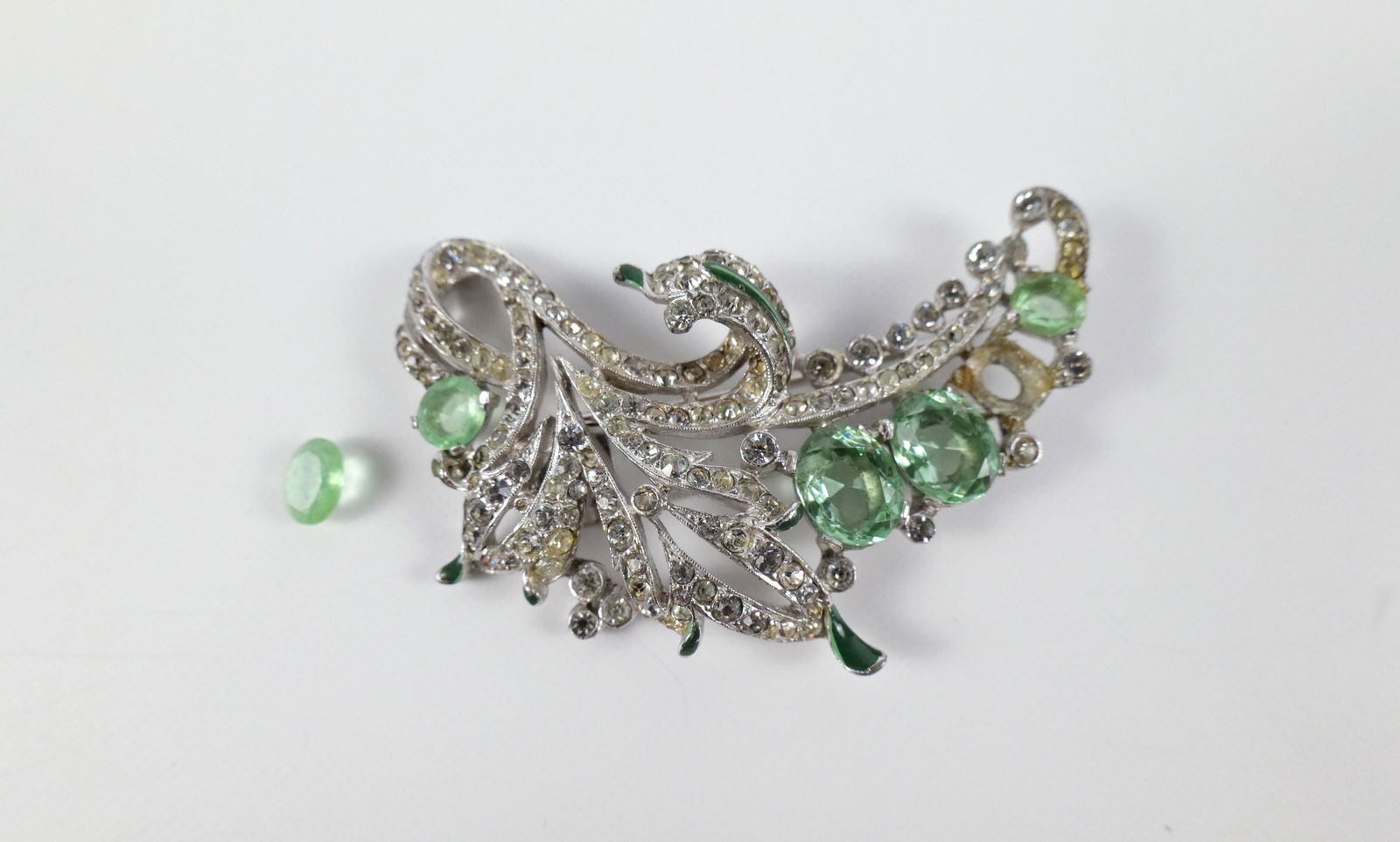 Vintage 1940s Rhinestone Fur Clip by Trifari: Vintage 1940s Rhinestone Fur Clip by Trifari. The lot includes a vintage 1940s 2.75'' x 1.77'' silver toned fur clip encrusted with white crystal rhinestones, oval pale green glass gems, and green ena