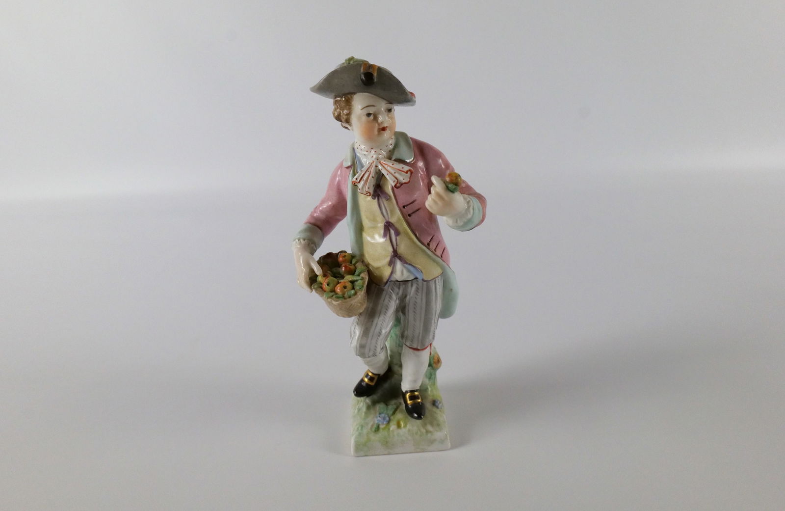 Antique KPM Porcelain Boy Figurine: Antique KPM Porcelain Boy Figurine. Hand Painted. Depicting boy holding basket of fruit. Small repair to coat tail, repair to arm, and some chips to leaves in basket. Measures 6" tall. All invoices mu