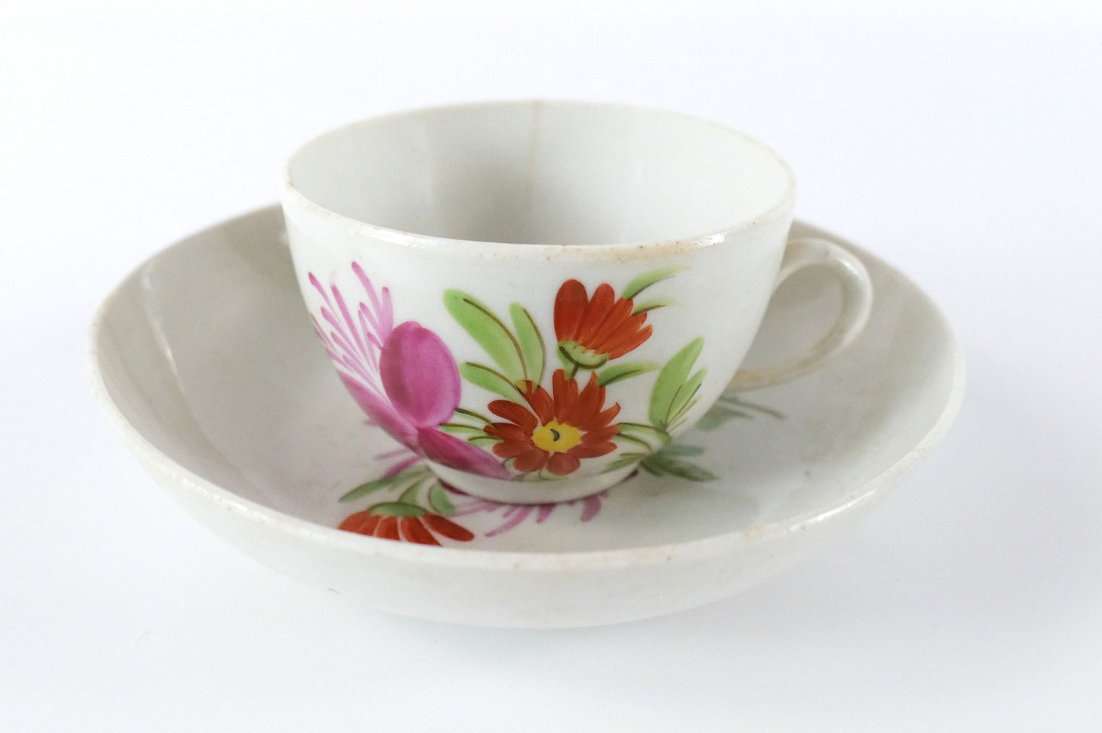 18th Century Hand Painted Porcelain Tea Cup and Saucer: 18th Century Hand Painted Porcelain Tea Cup and Saucer. Has floral motif. The saucer has 2 hairline cracks by the edge. The cup has one hairline crack, a firing discoloration spot, and some flea bite