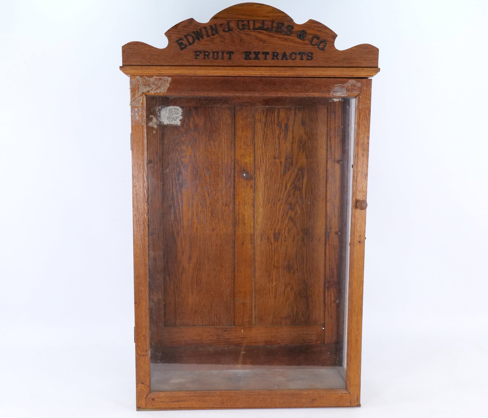 Antique Edwin J. Gillies and Co. Fruit Extracts Oak Store Display Cabinet: An antique retail display cabinet constructed from oak with a glazed front and side panels. The cabinet features a decorative arched and stepped pediment with black stenciled lettering that reads: