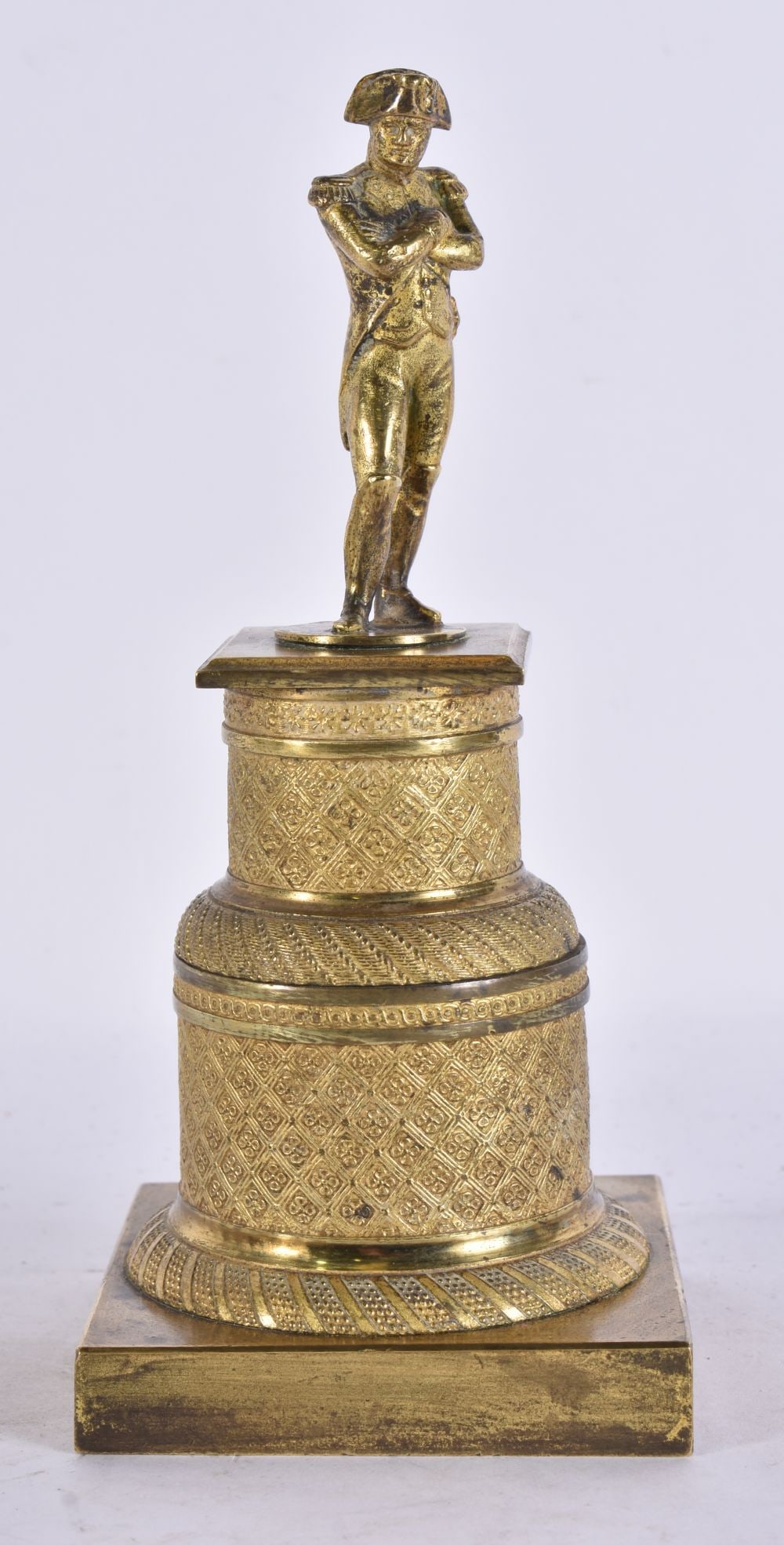 AN ANTIQUE FRENCH GRAND TOUR GILT BRONZE FIGURE OF NAPOLEON standing with arms folded in uniform wea: AN ANTIQUE FRENCH GRAND TOUR GILT BRONZE FIGURE OF NAPOLEON standing with arms folded in uniform wearing a bicorne hat mounted on a tiered cylindrical plinth with engine turned diaper decoration and b