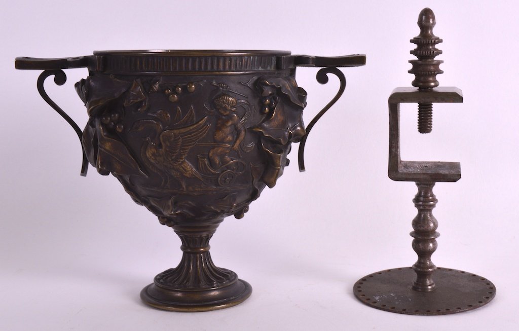 A 19TH CENTURY EUROPEAN BRONZE TWIN HANDLED CUP after a: A 19TH CENTURY EUROPEAN BRONZE TWIN HANDLED CUP after a Roman Skyphos model, decorated with classical scenes, together with a polished steel clamp. 5.75ins & 6.25ins high. (2) £50-75