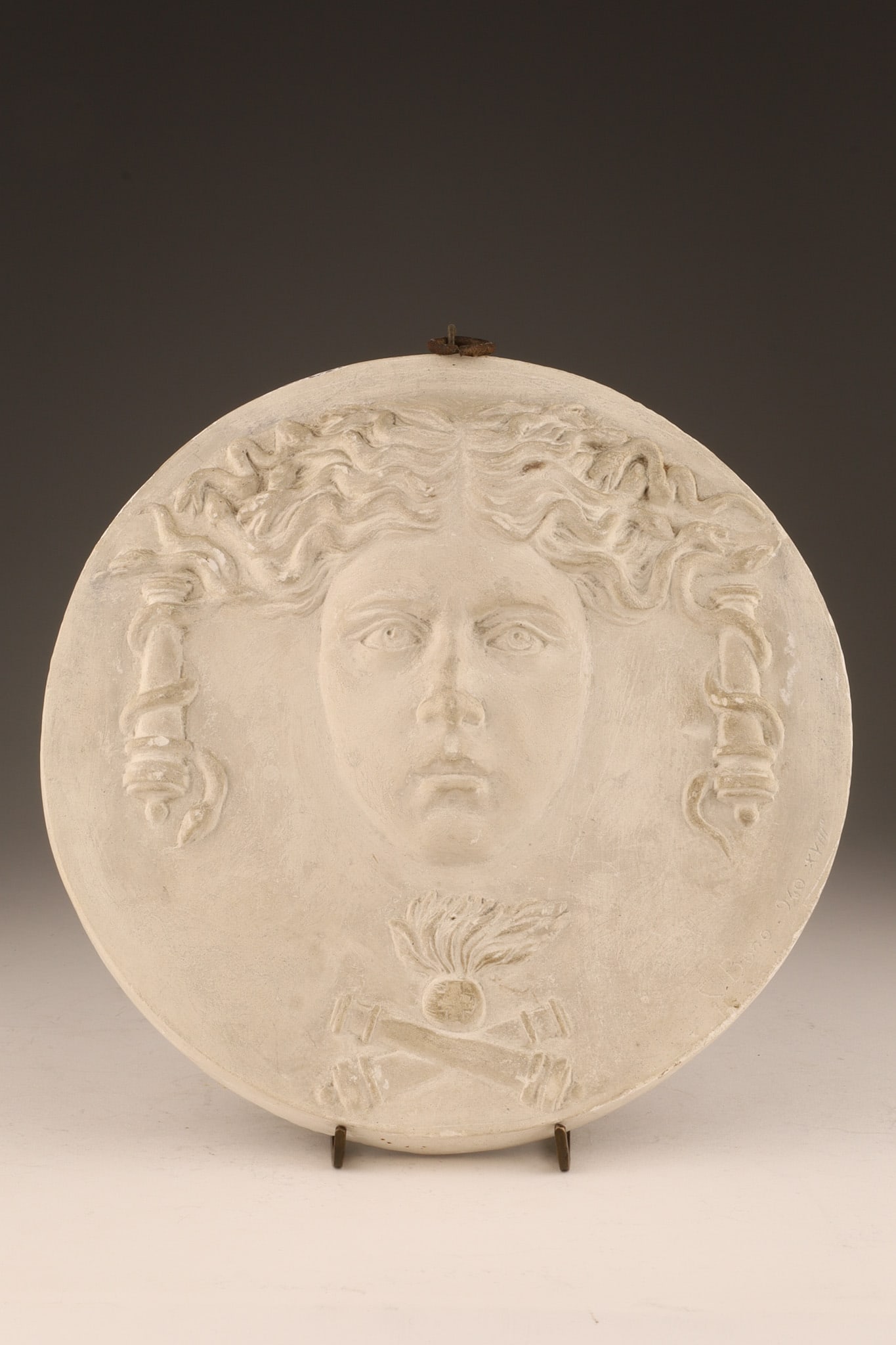 Neoclassical Medusa Plaster Medallion Signed Bono: Neoclassical Medusa Plaster Low Relief Medallion Signed C. Bono 940 XVIII. Face of Medusa surrounded by cannons above crossed cannons below a flaming pot. 8.5 inches diameter.
