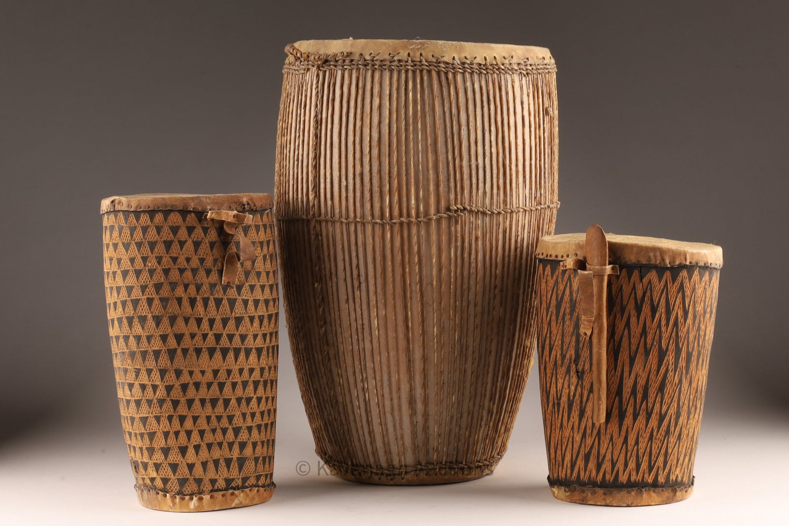 Collection of African Drums: Collection of African Drums, two smaller geometric incised pattern drums, one string bound drum all with hide skins. Taller measures 14 inches, smaller measure 9 inches.