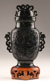 Chinese Spinach Jade Vase and Cover: Chinese Spinach Jade Vase and Cover, body carved with stylized taotie masks and dragon-shaped handles. Loss to lid on one side, mounted to shaped wood stand, measuring 7 inches H with stand, vase meas