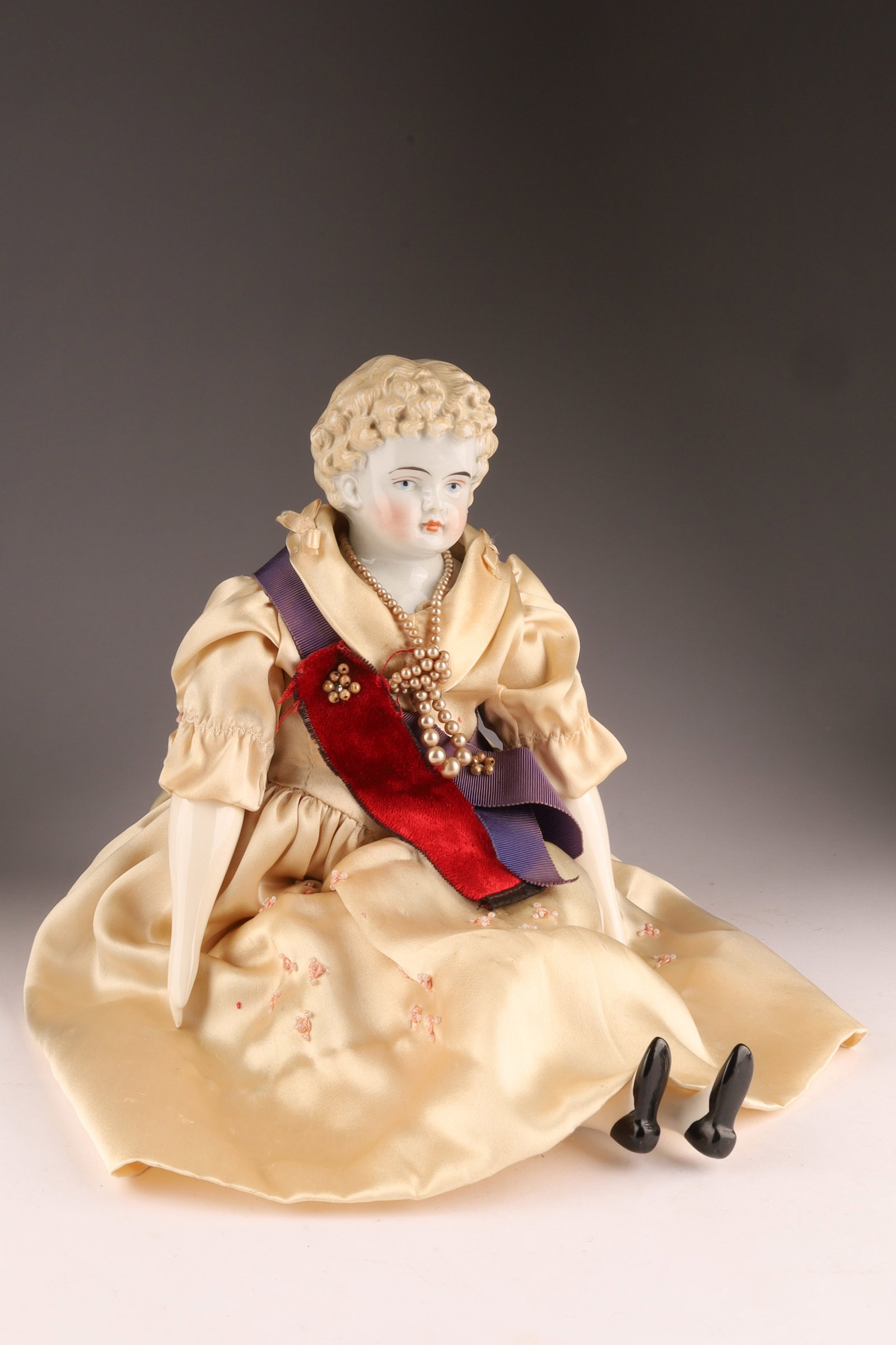 Antique Porcelain Doll in Formal Attire, Blonde Hair Blue Eyes: Antique porcelain doll dressed in formal attire, featuring molded blonde curly hair, blue eyes and rosy cheeks. The doll has a glazed porcelain upper torso, hands, and legs with a soft body cov