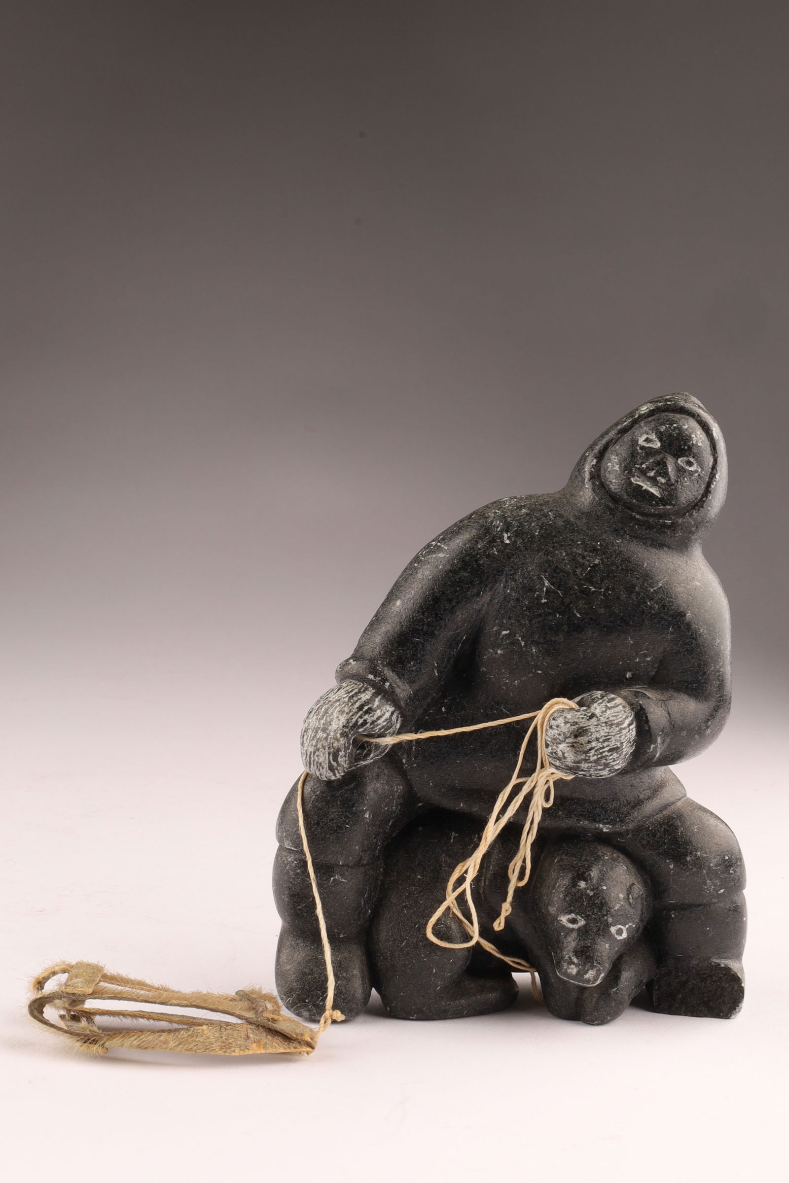 Andrew Valik Inuit Soapstone Hunter with Bear and Sled Signed Sculpture, 5 Inches: Carved black soapstone figure depicting an Inuit hunter in traditional parka seated above a bear, holding a line attached to a miniature sled. The figure is executed in a compact, rounded form with in